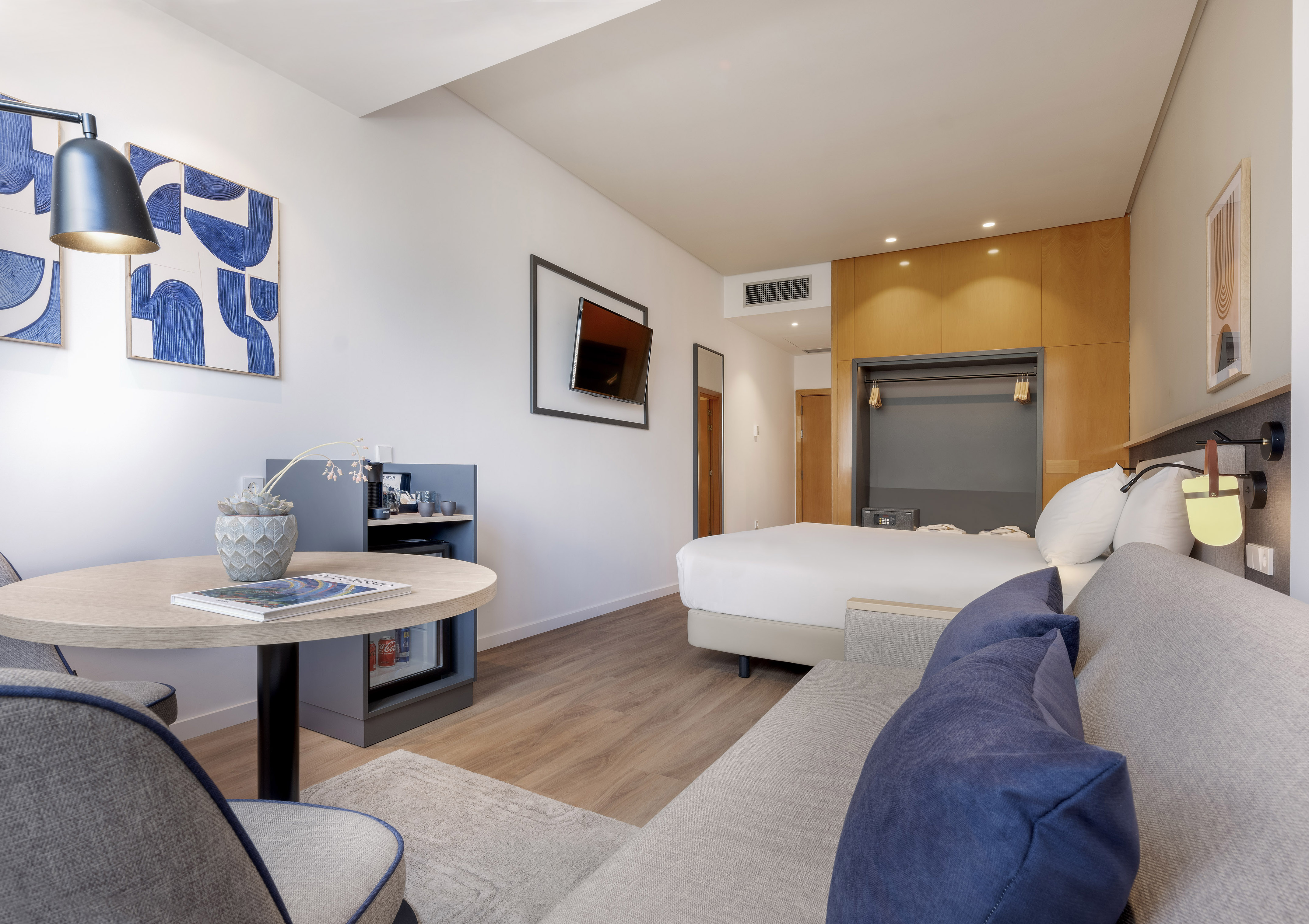 Modern hotel room with bed, grey sofa, round table, TV, and blue art.