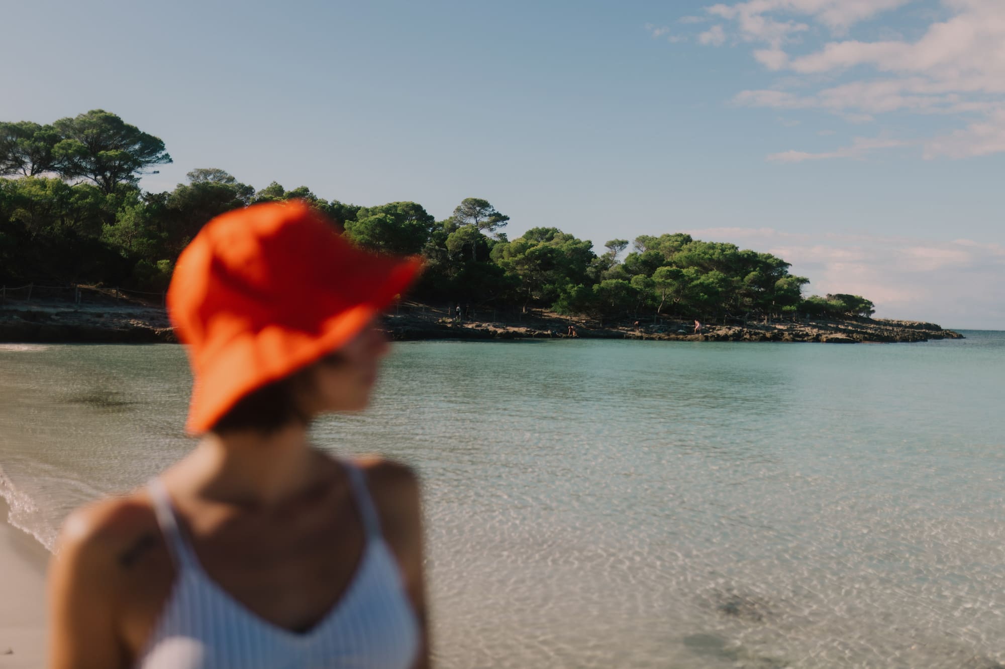 Mujer con sombrero naranja en cala de aguas turquesas y pinos.
