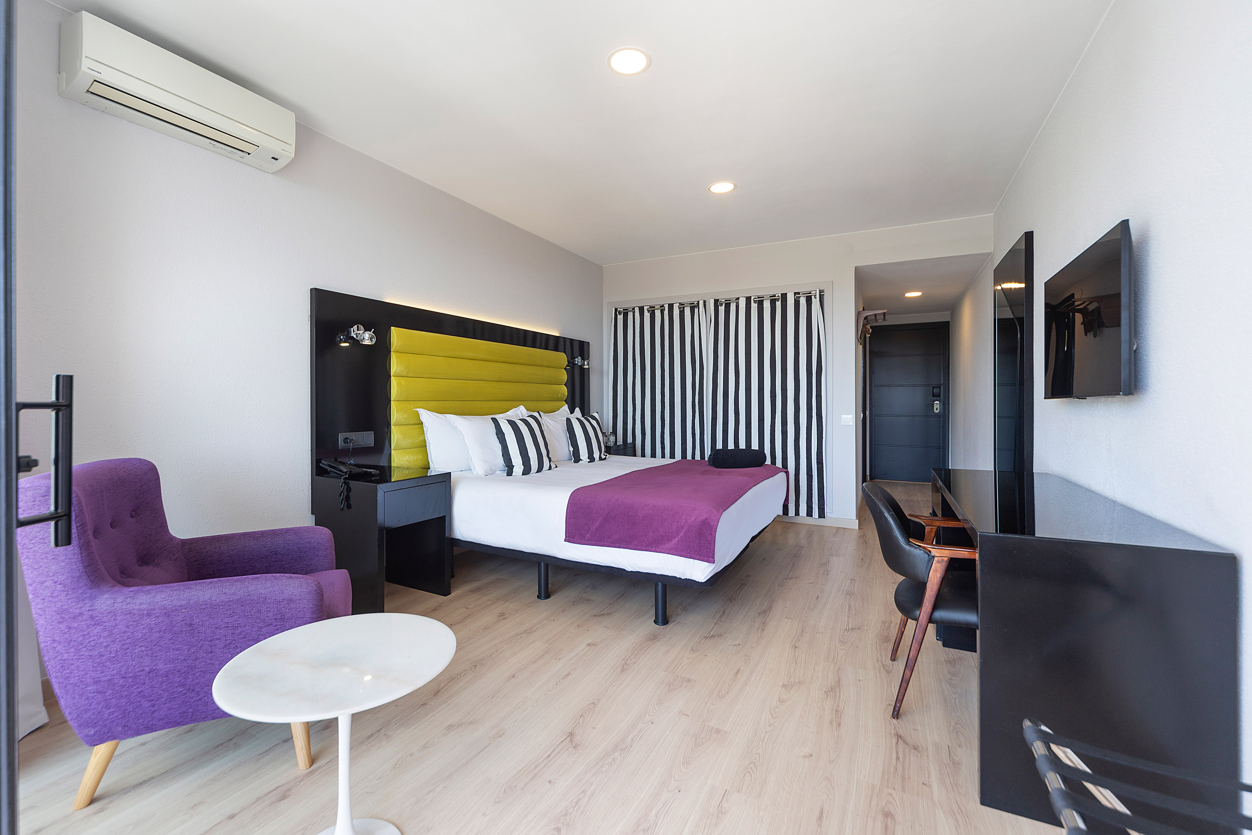 Vibrant hotel room with yellow headboard, purple accents, and light wood flooring.