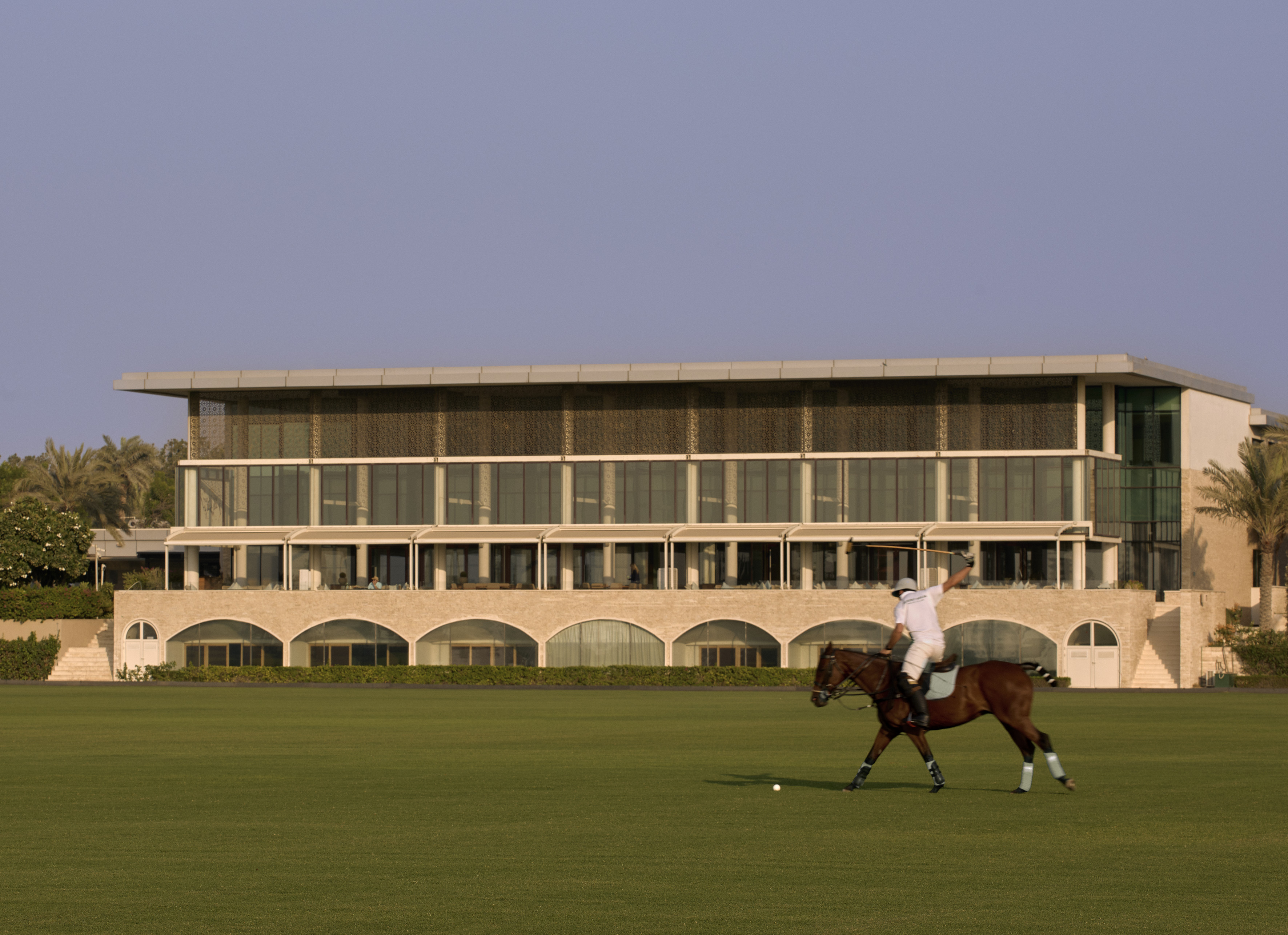 Polo player on horse on a green field, modern building with arched base.