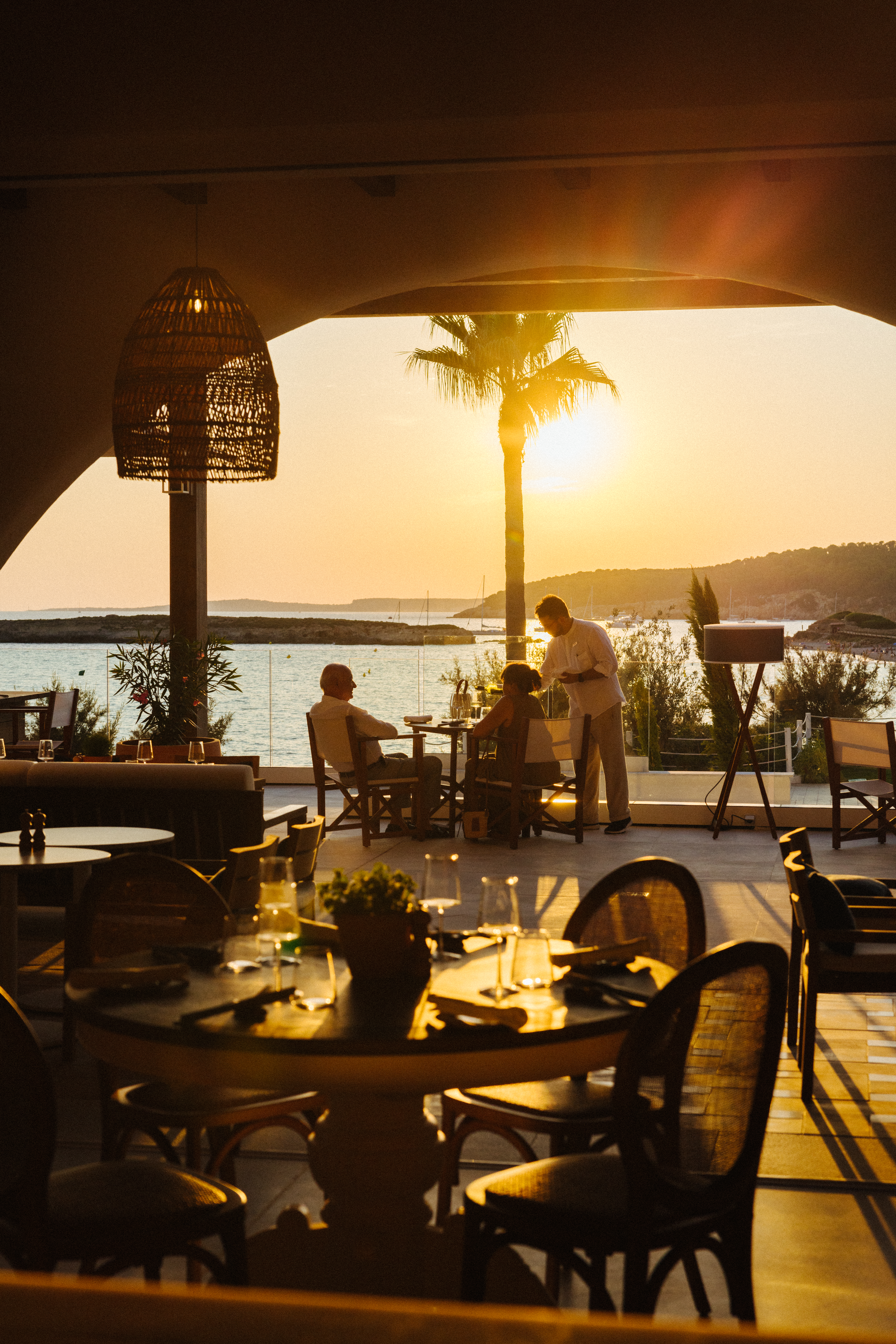 Oceanfront restaurant terrace, golden sunset, waiter serving diners.
