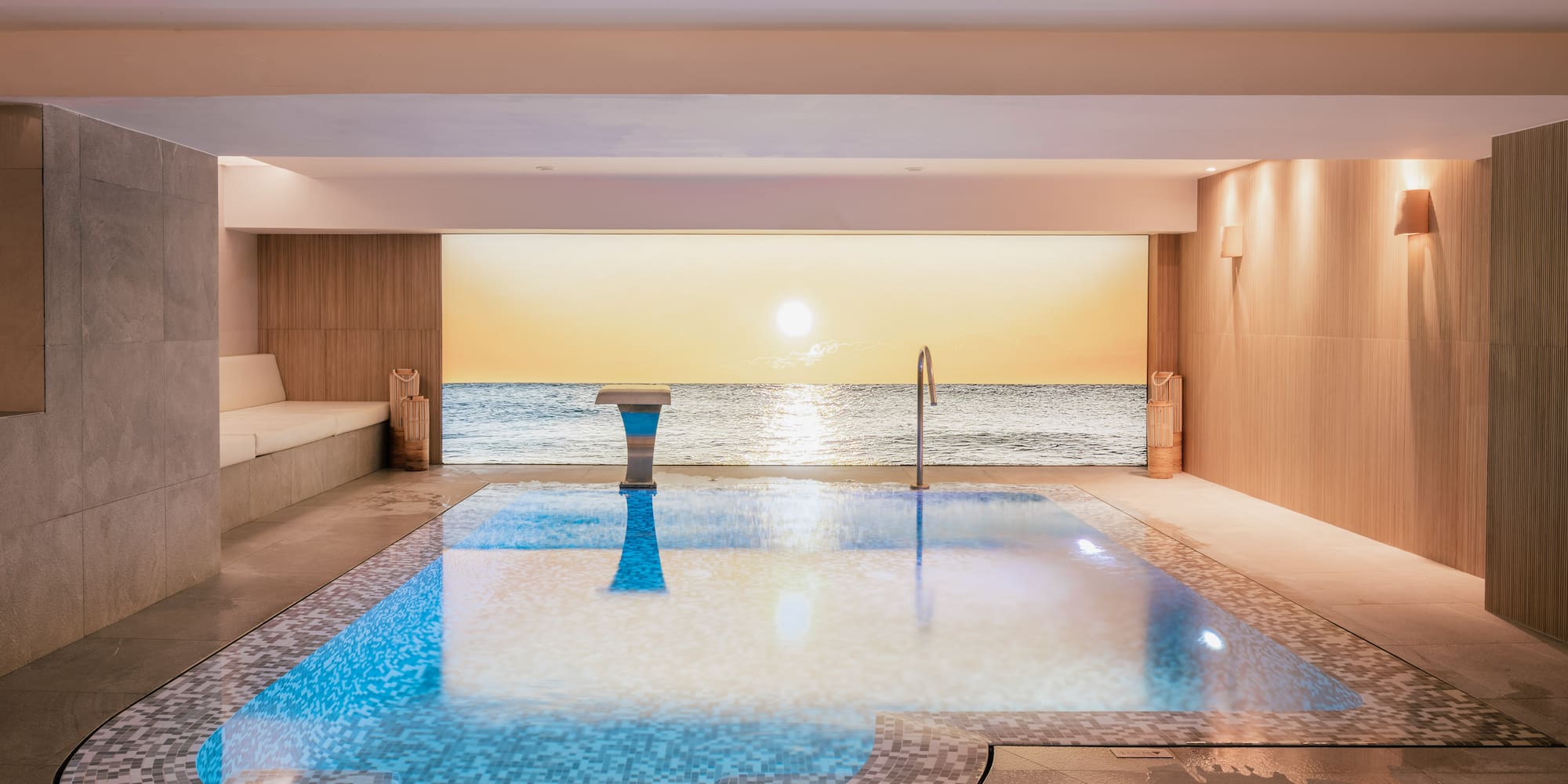 Indoor pool with mosaic tiles, ocean sunset view, luxury spa design.