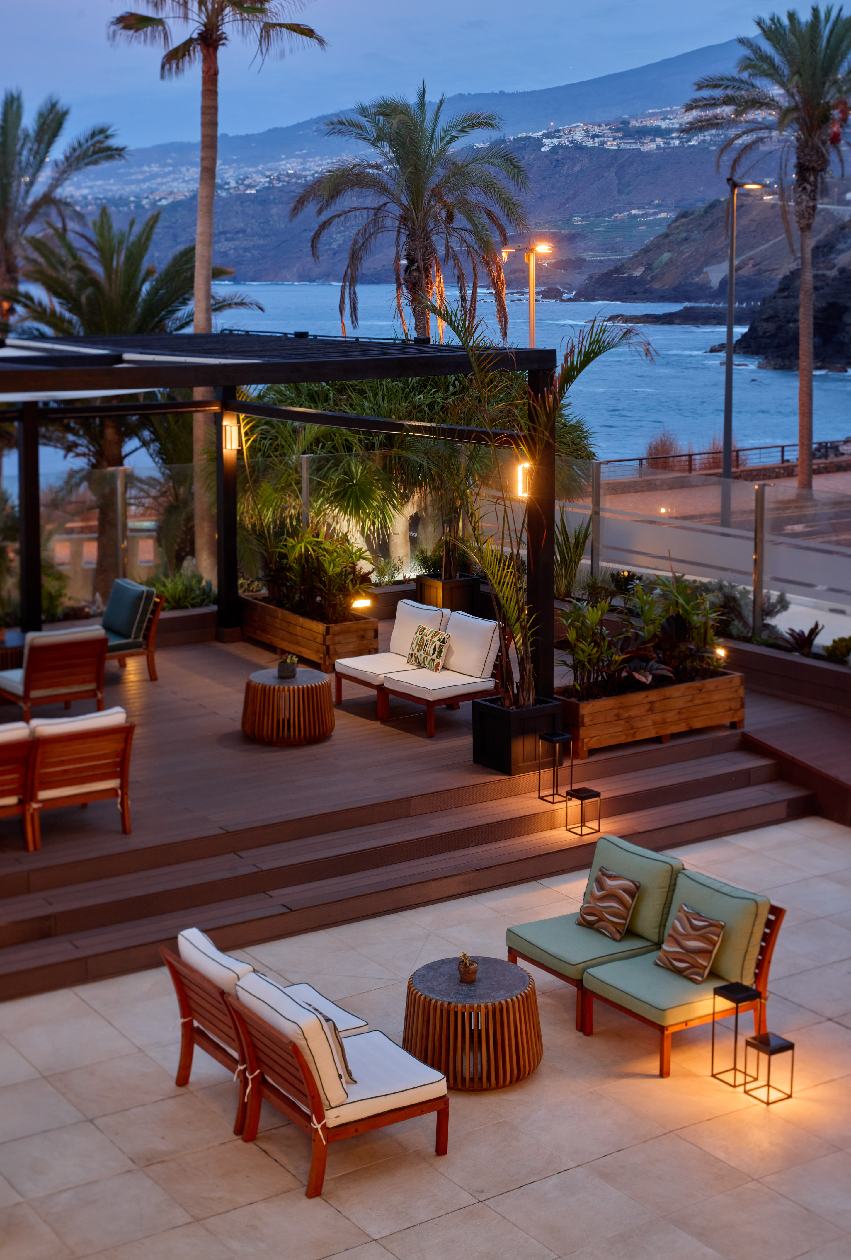Outdoor lounge patio with ocean view at dusk, palm trees, modern furniture.