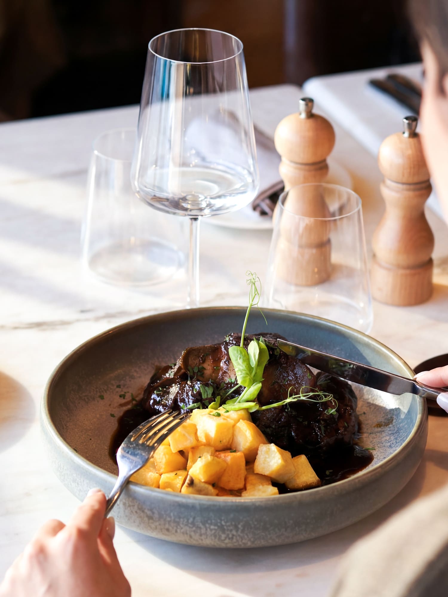 Person cutting braised meat with potato cubes at a restaurant table.
