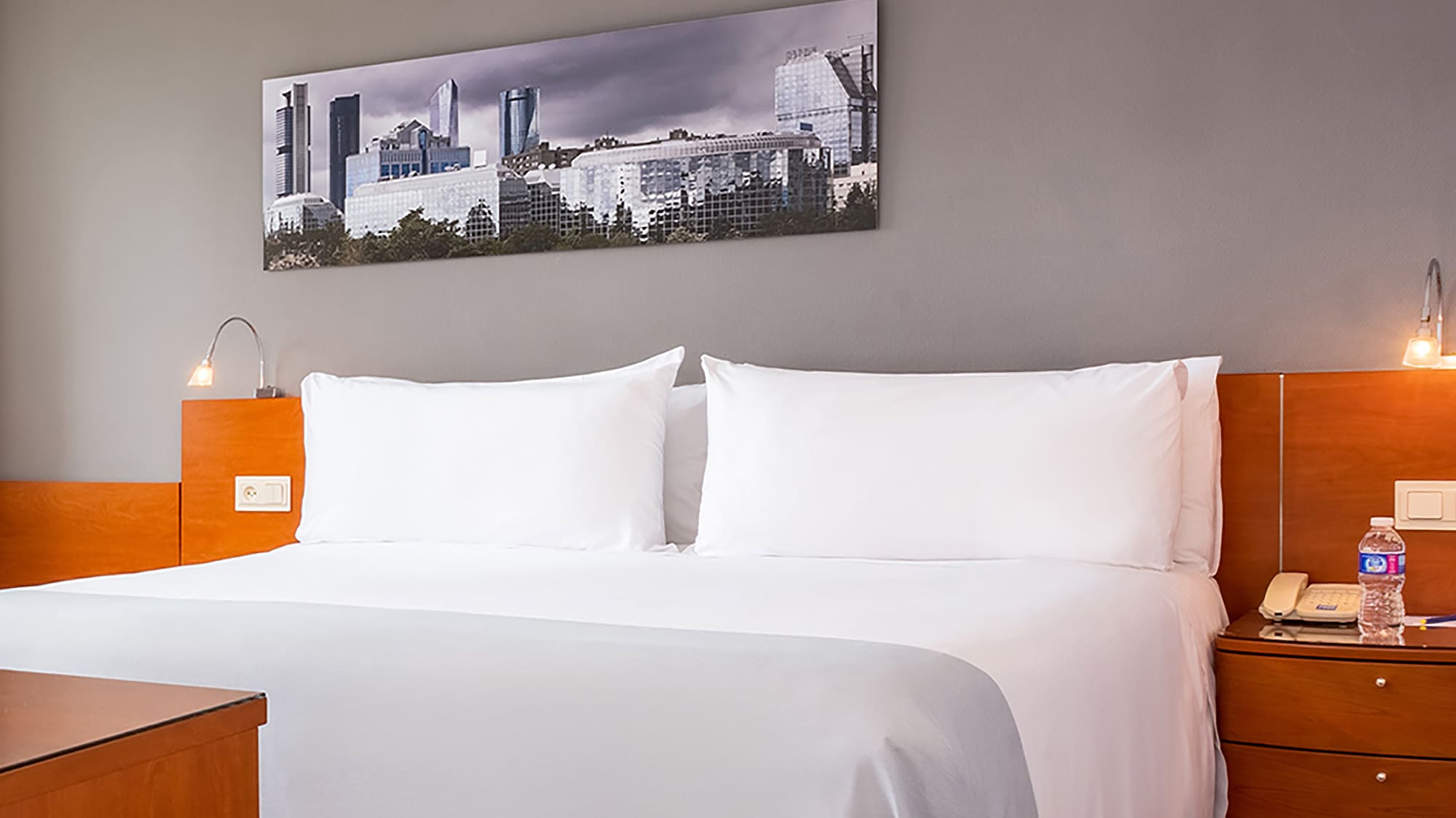 Modern hotel room with white bed, wooden floor, and cityscape artwork.