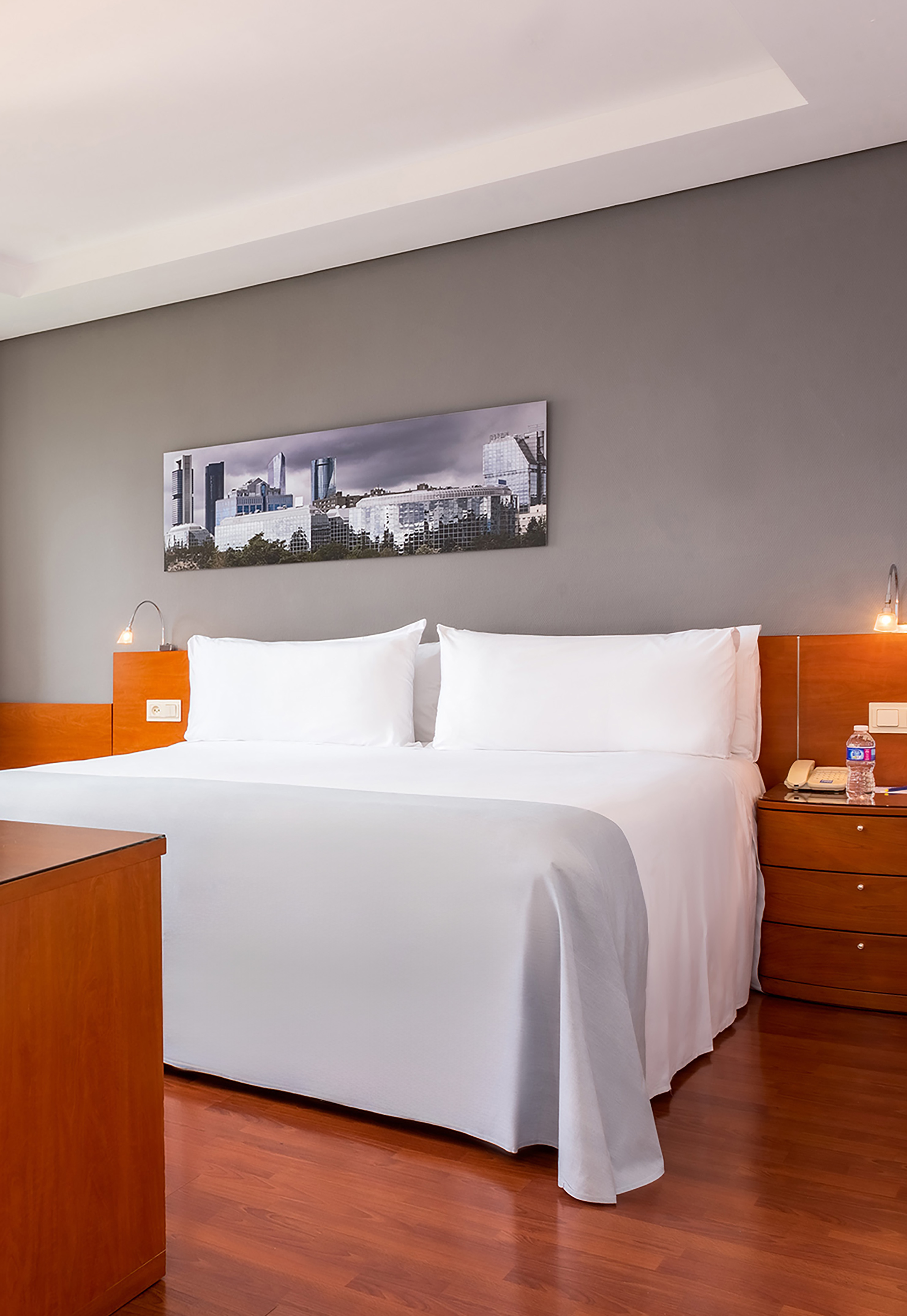 Modern hotel room with white bed, wooden floor, and cityscape artwork.