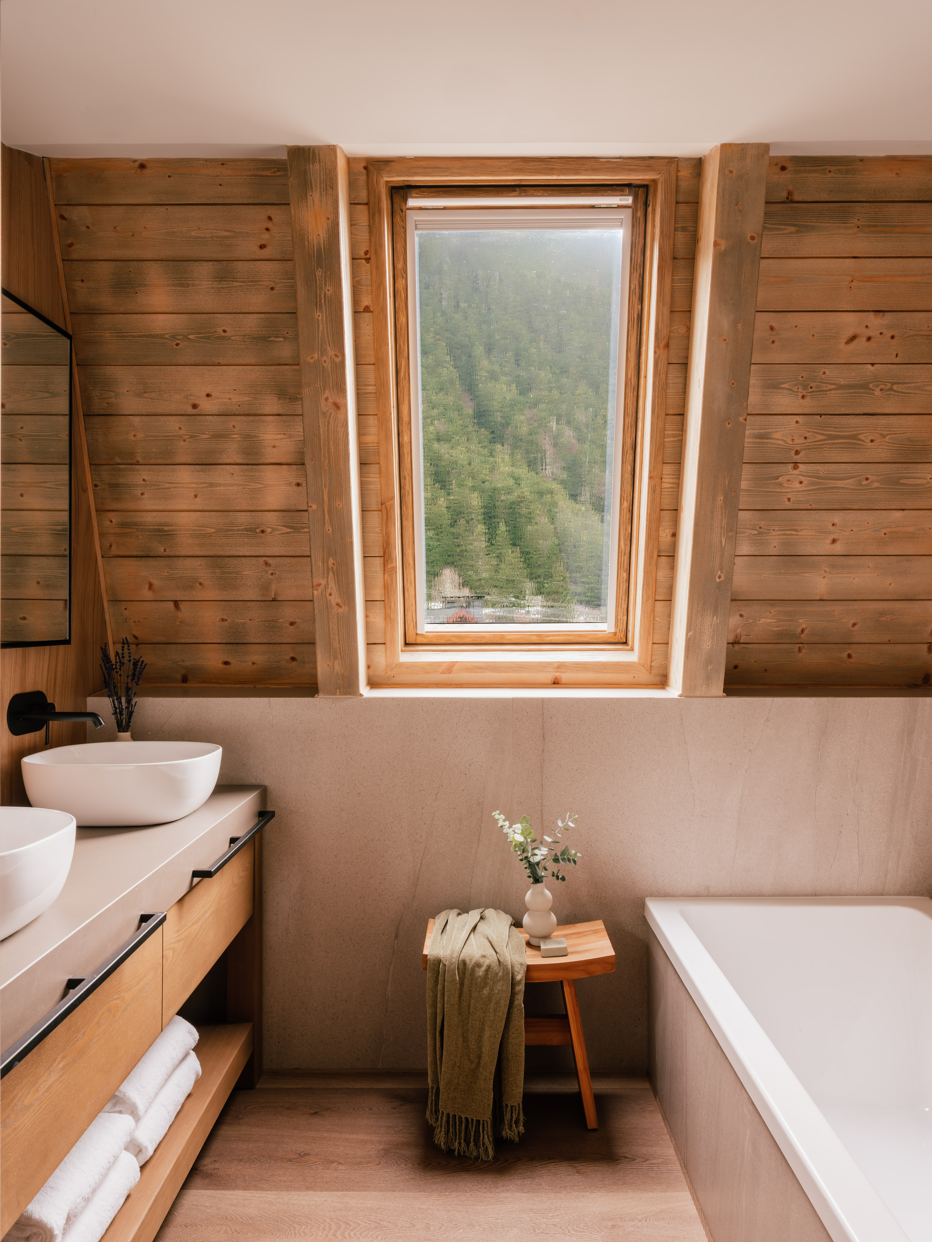 Rustic wood paneled bathroom with forest view, double vanity, and bathtub.