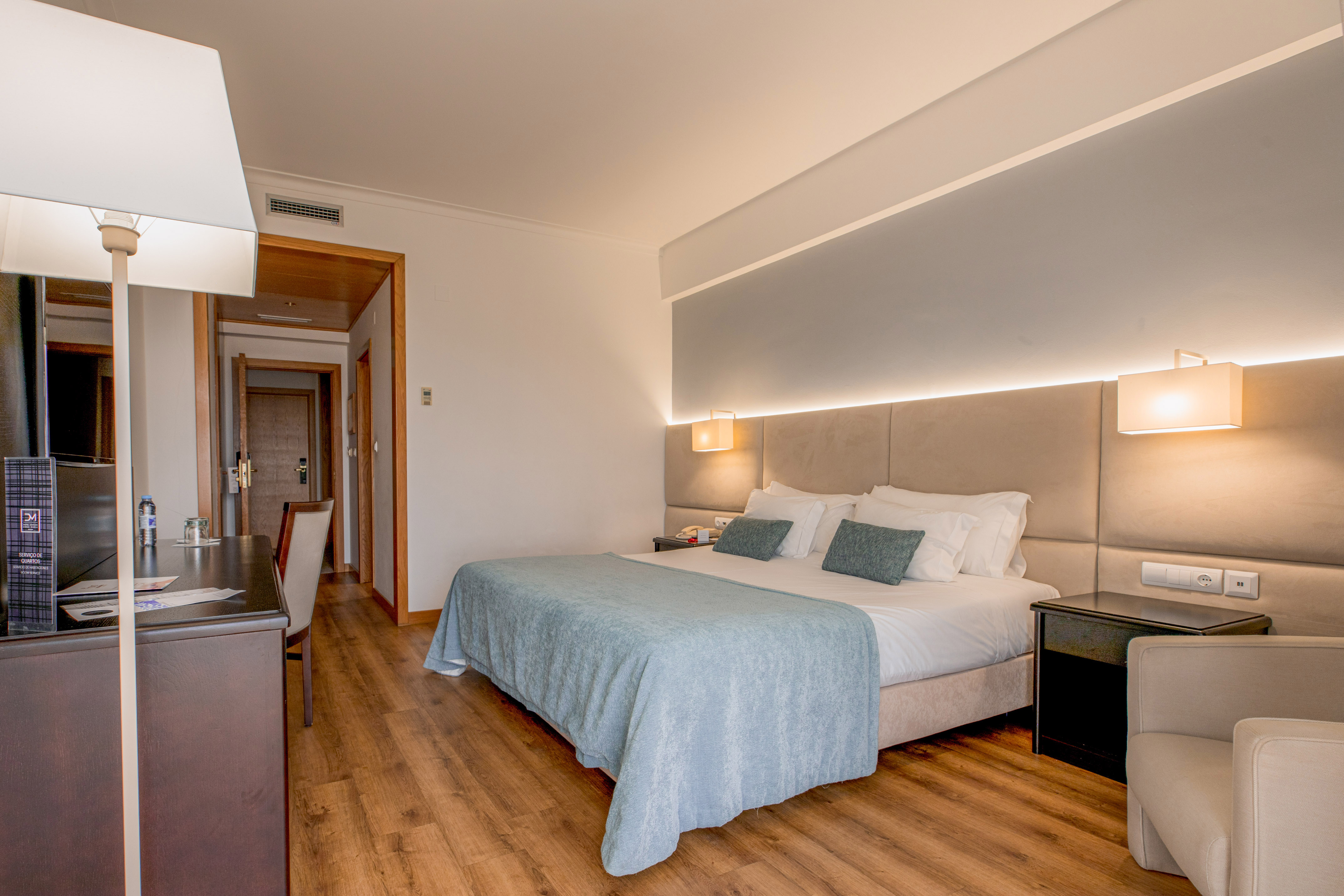 Modern hotel room with king bed, light blue blanket, wooden floor, and desk.