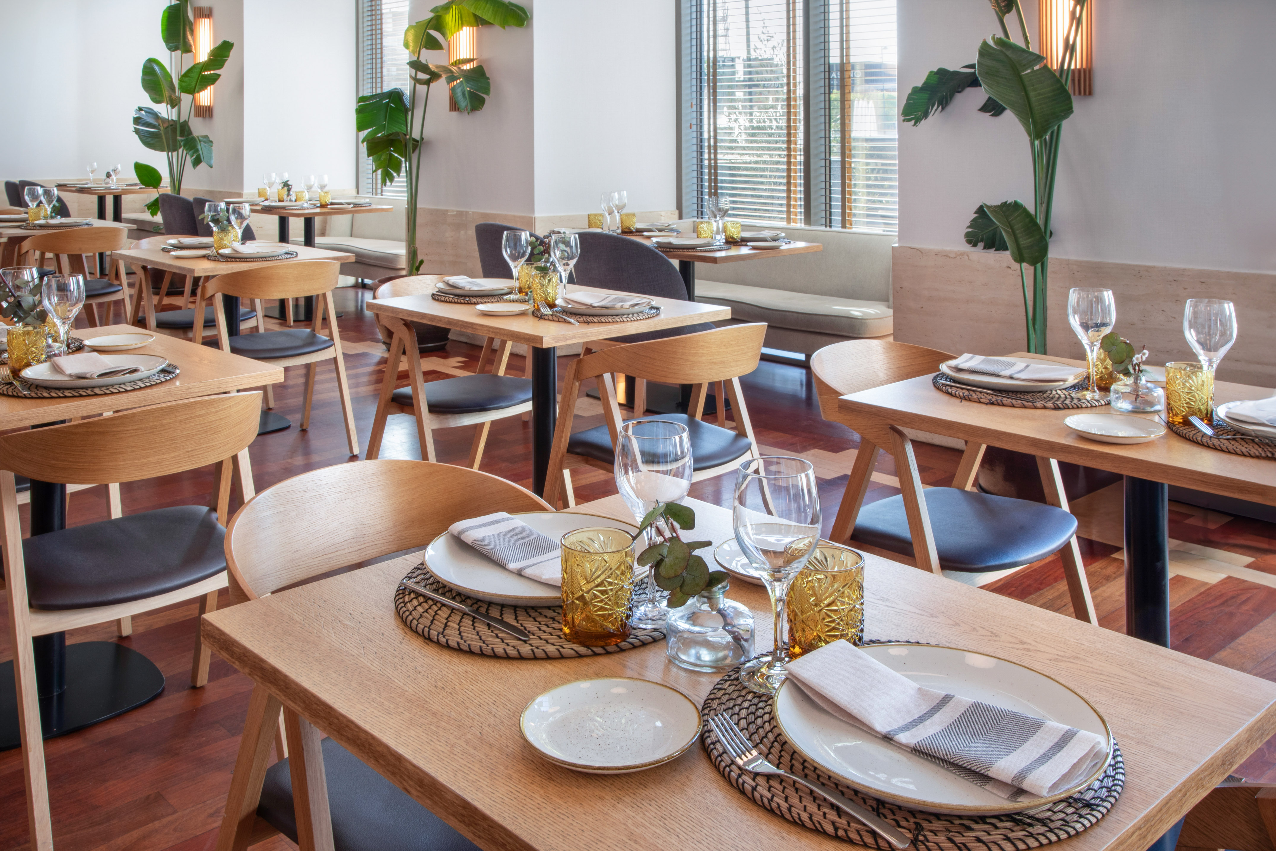 Modern restaurant dining room with wooden tables, place settings, and tropical plants.