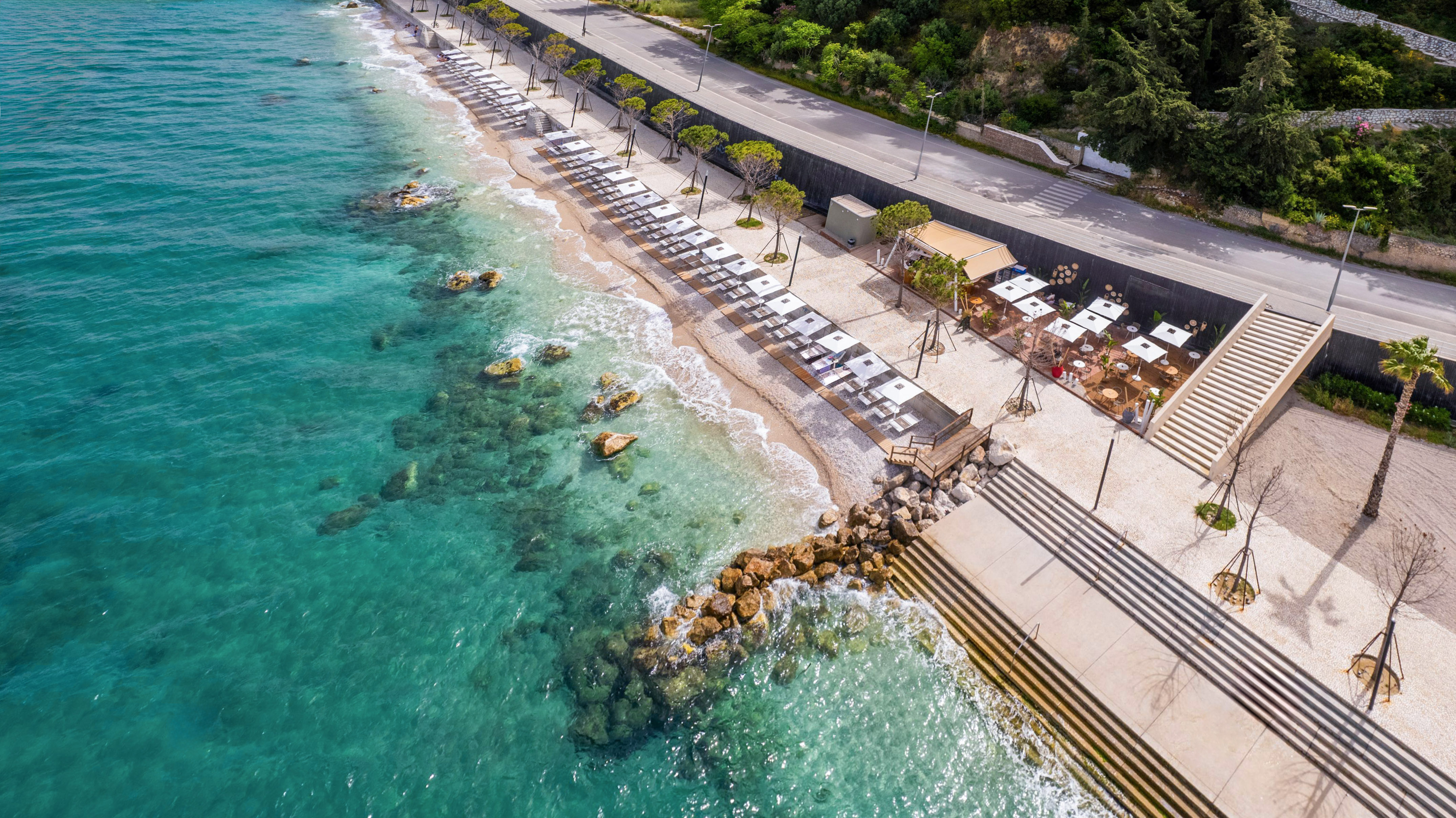 Aerial view of modern beach with turquoise sea, lounge chairs, cafe, and coastal road.