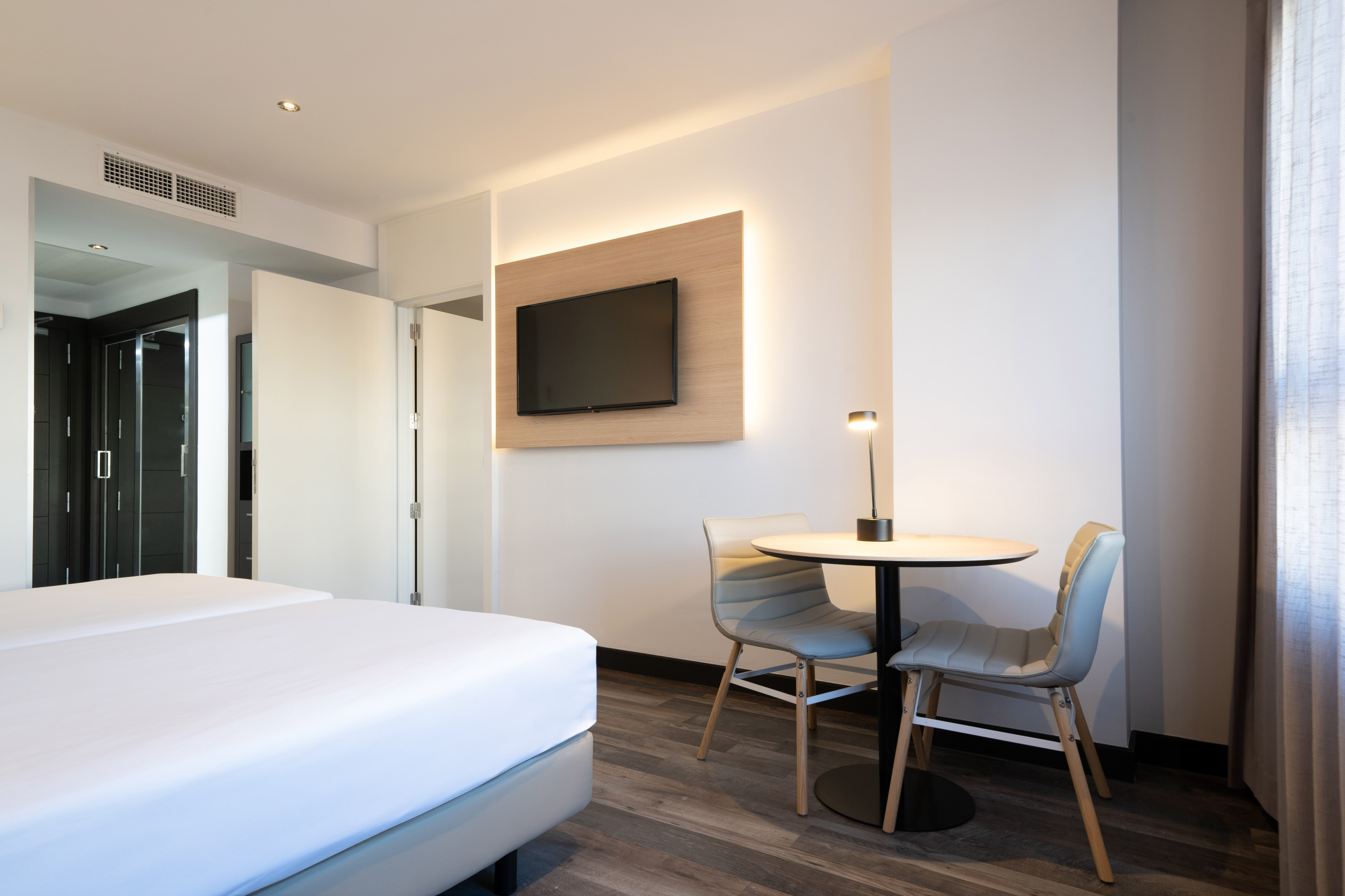 Modern hotel room with twin beds, wall-mounted TV, table, and two chairs.