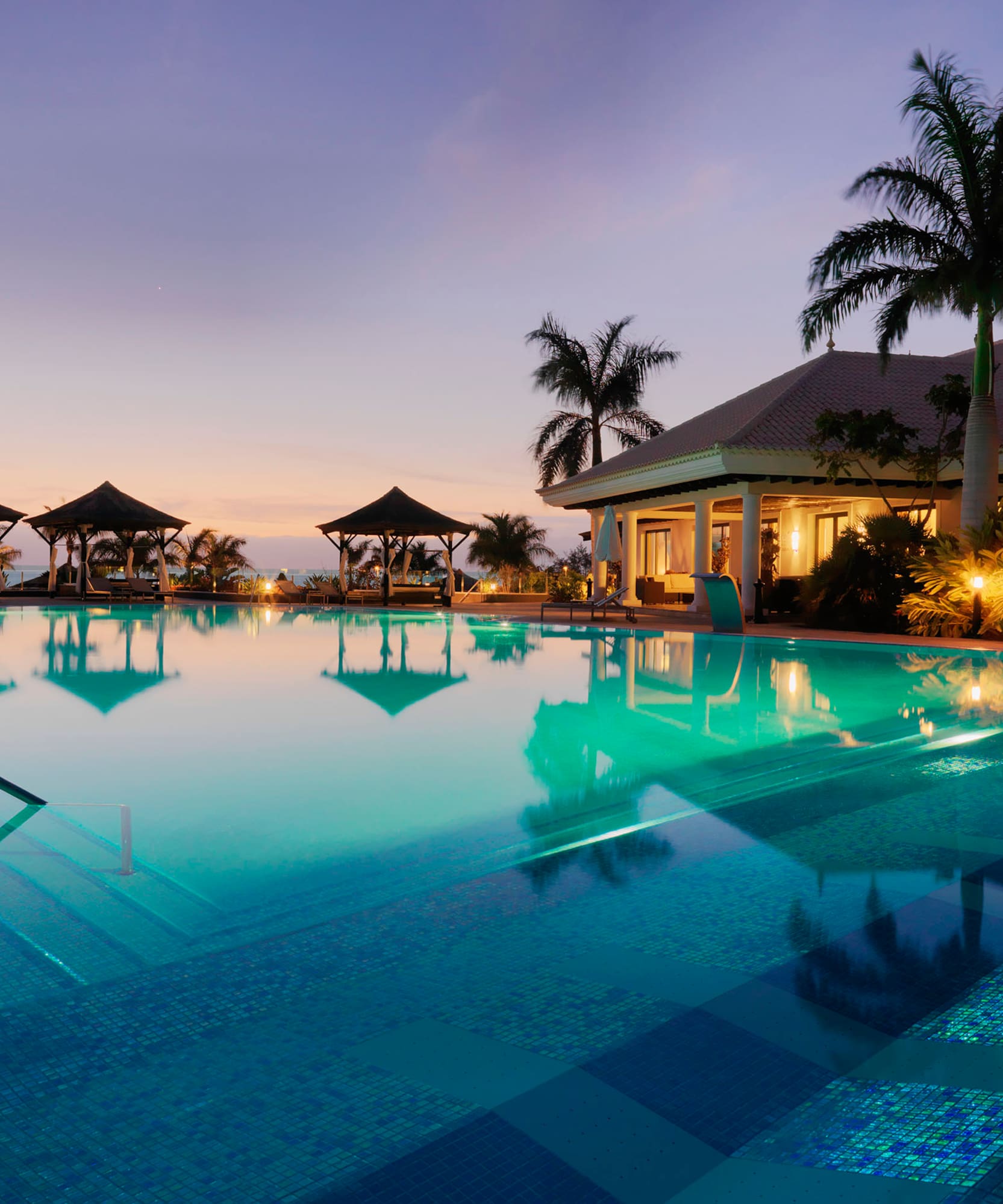 Resort de lujo al anochecer con piscina, palmeras y cenadores.