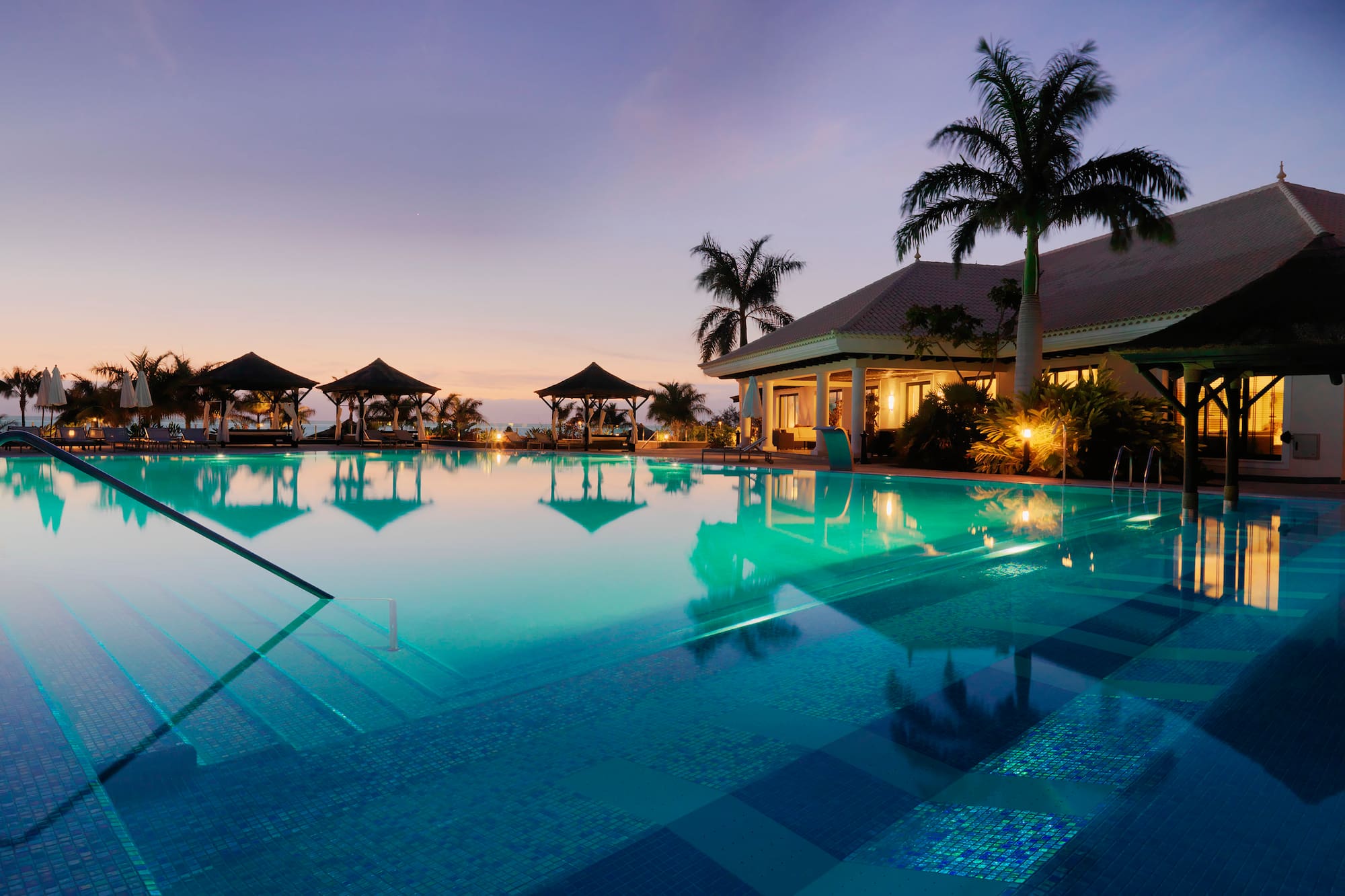 Resort de lujo al anochecer con piscina, palmeras y cenadores.