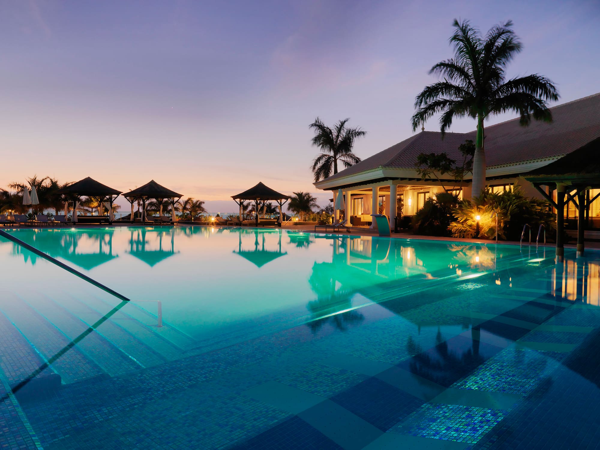 Resort de lujo al anochecer con piscina, palmeras y cenadores.