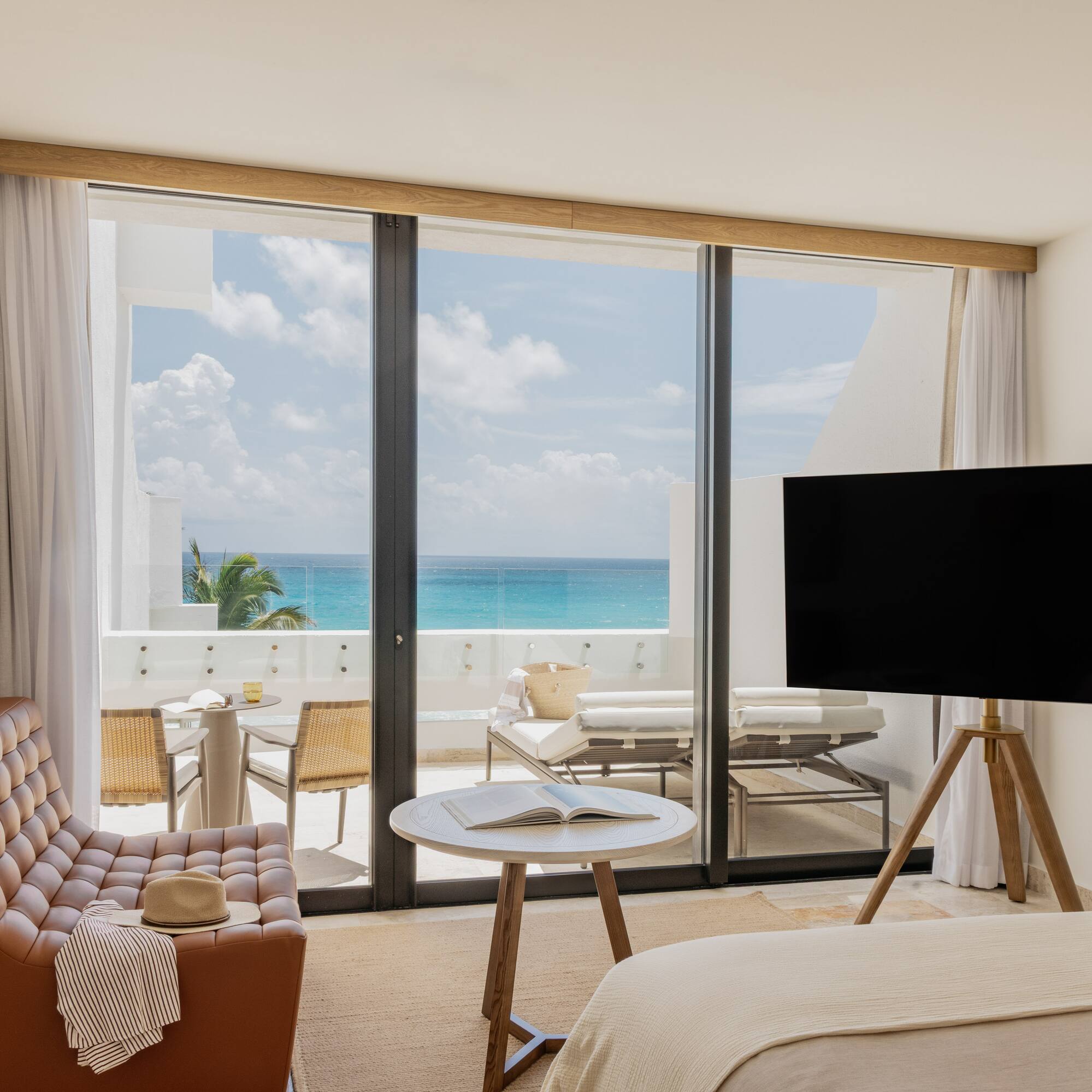 Modern hotel room with turquoise ocean view balcony, armchair, and TV.