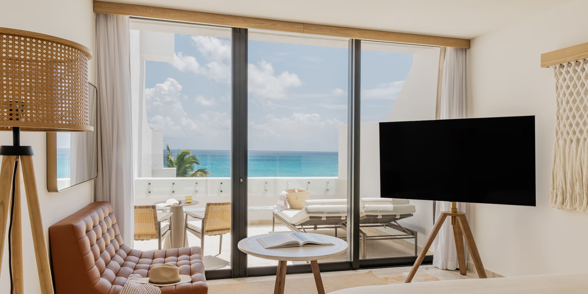 Modern hotel room with turquoise ocean view balcony, armchair, and TV.