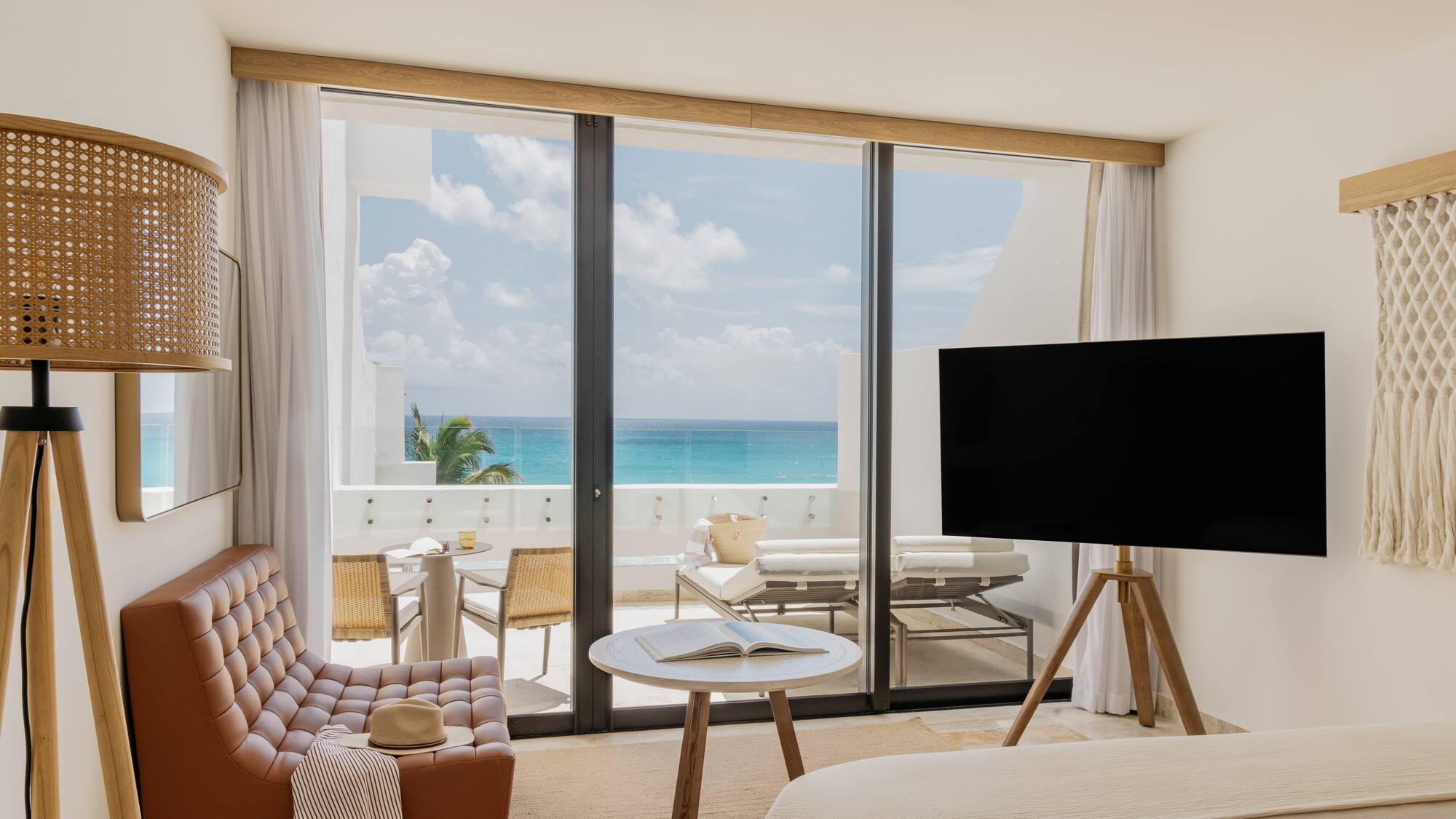 Modern hotel room with turquoise ocean view balcony, armchair, and TV.