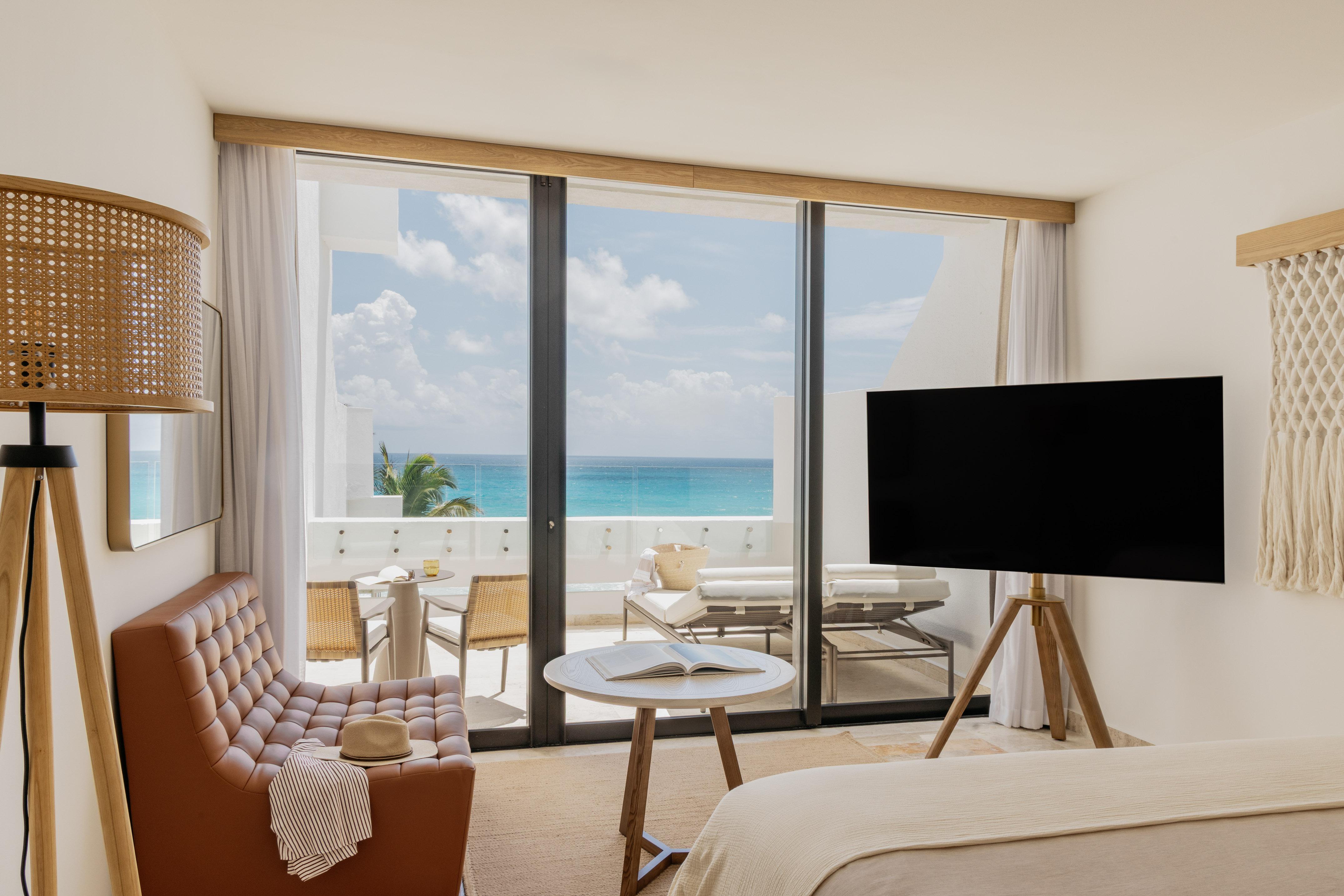 Modern hotel room with turquoise ocean view balcony, armchair, and TV.