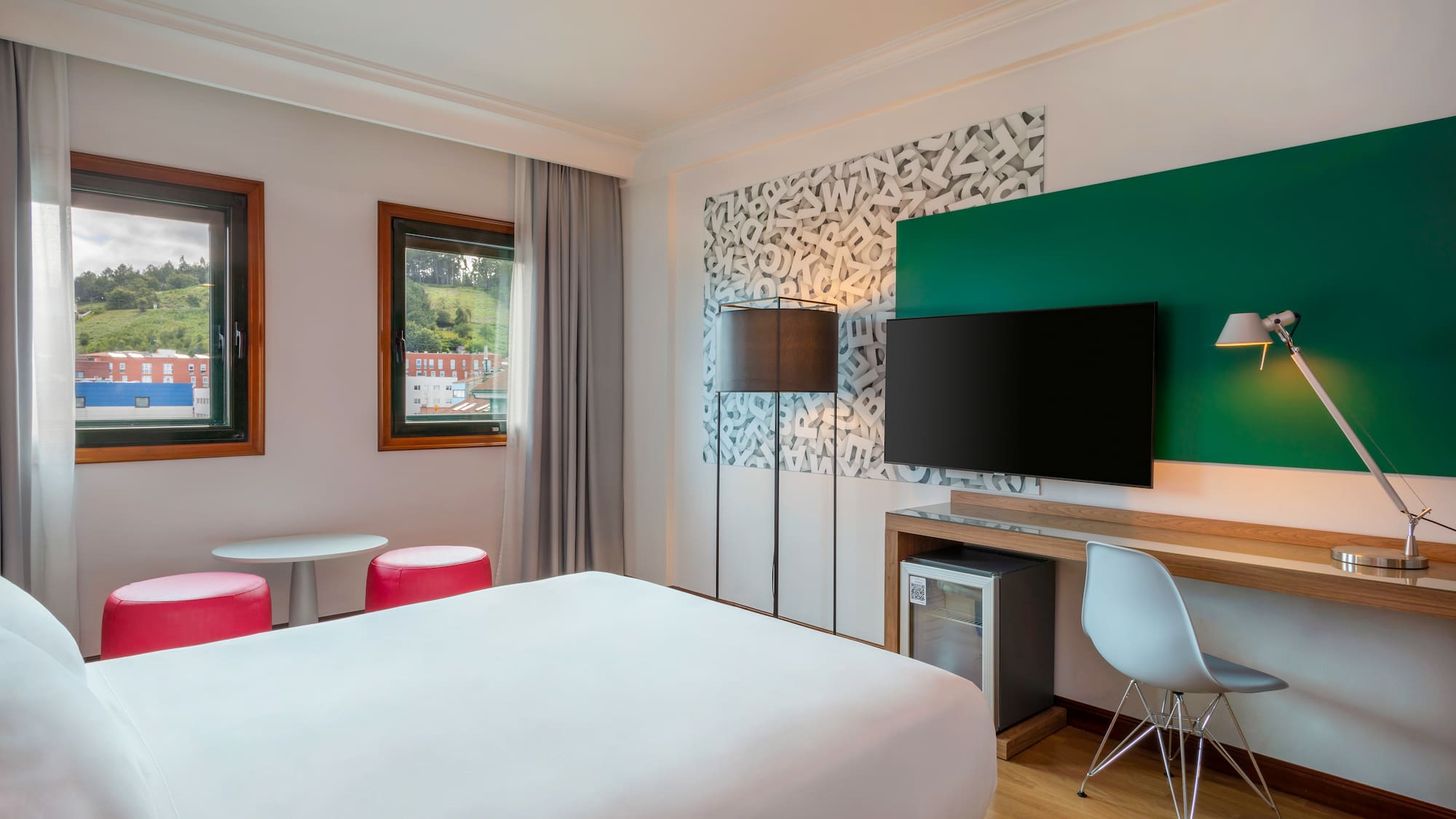 Bright hotel room with a comfortable bed, workspace, and view from two windows.