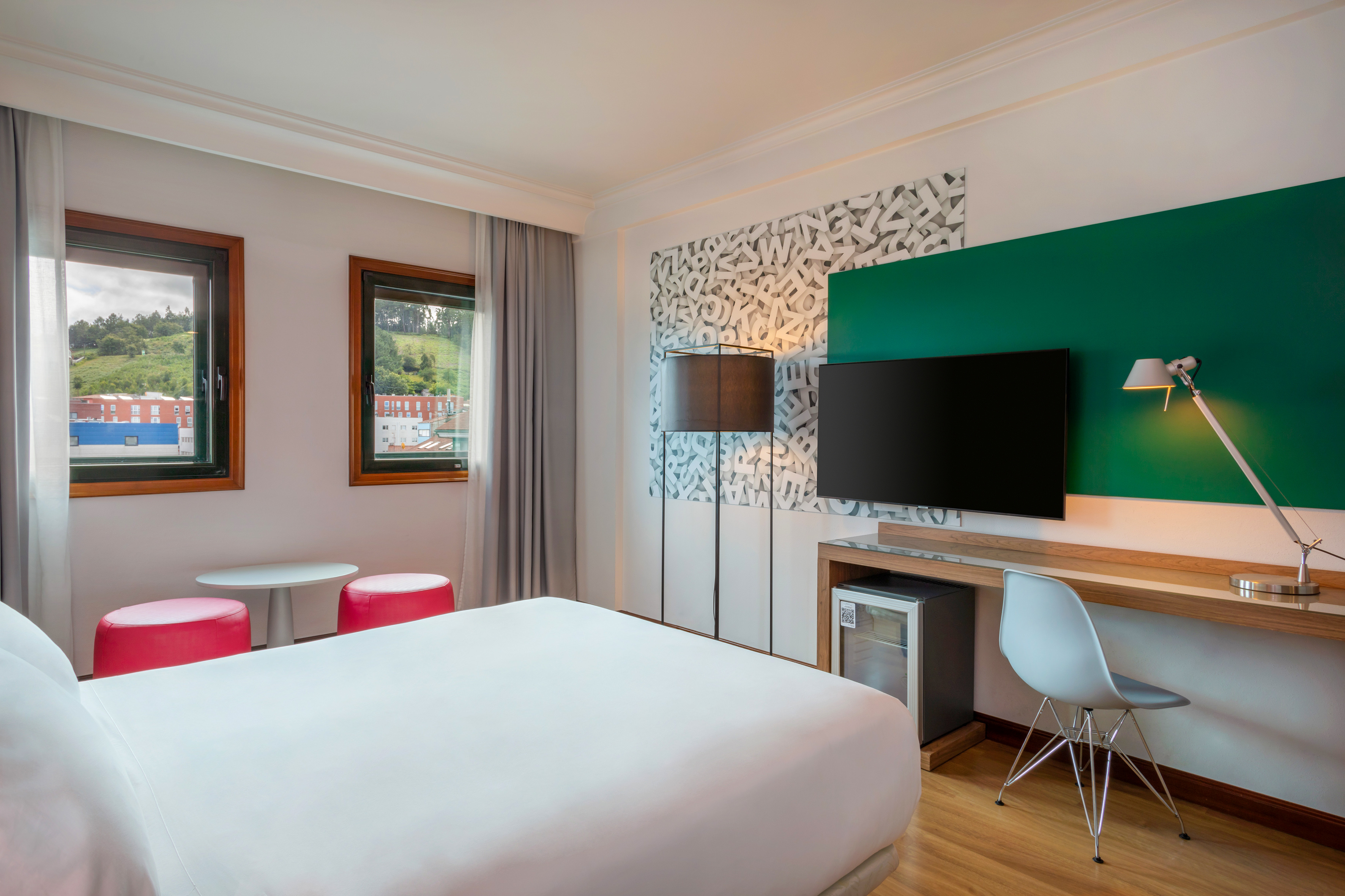 Bright hotel room with a comfortable bed, workspace, and view from two windows.