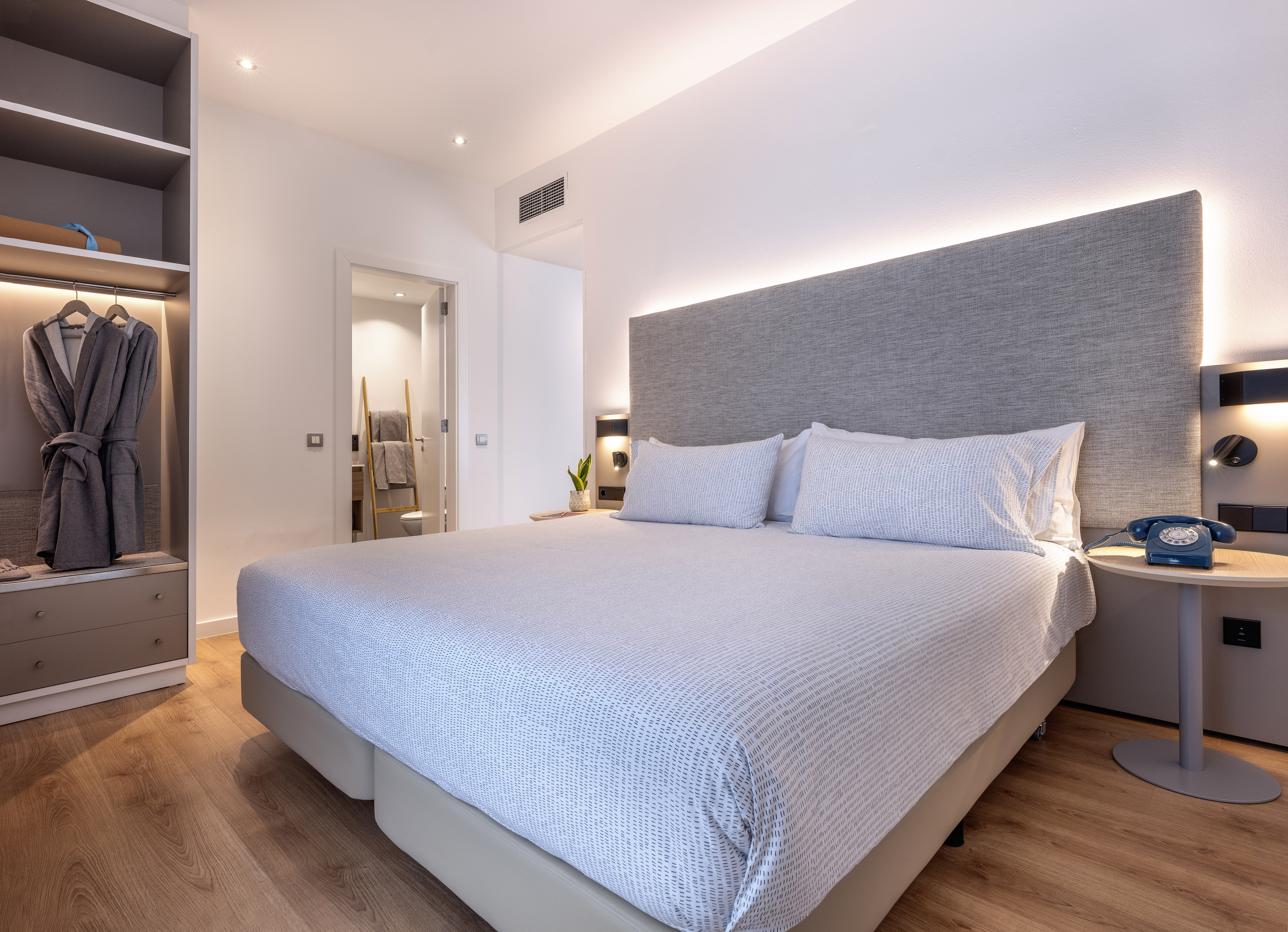 Modern hotel bedroom with large bed, grey headboard, and open wardrobe.