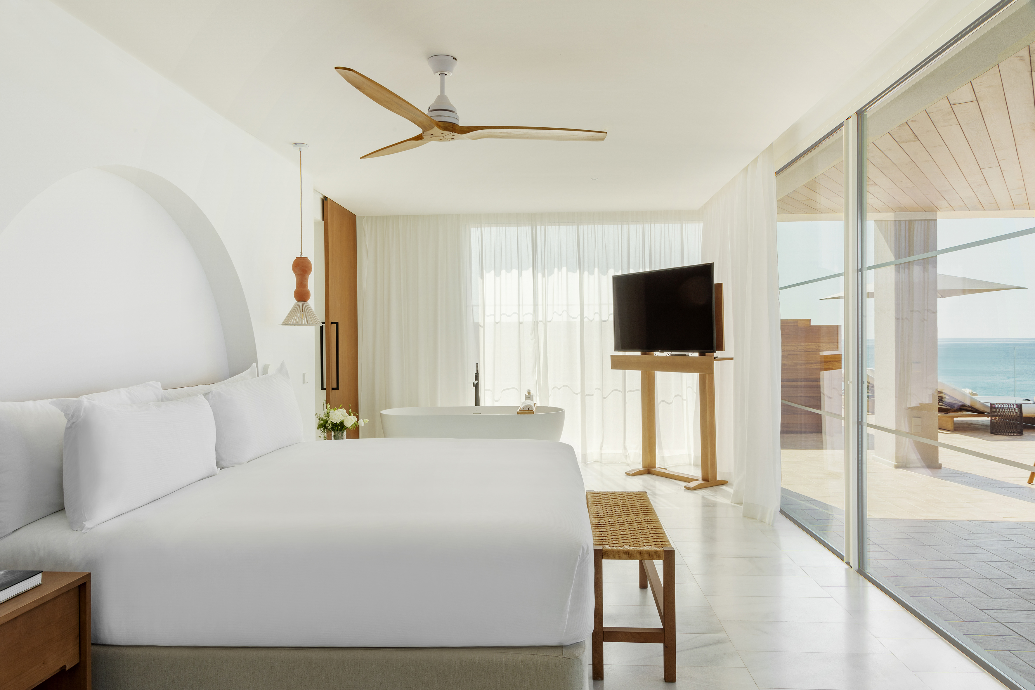 Luxury hotel bedroom with ocean view, white bed, freestanding tub, and fan.