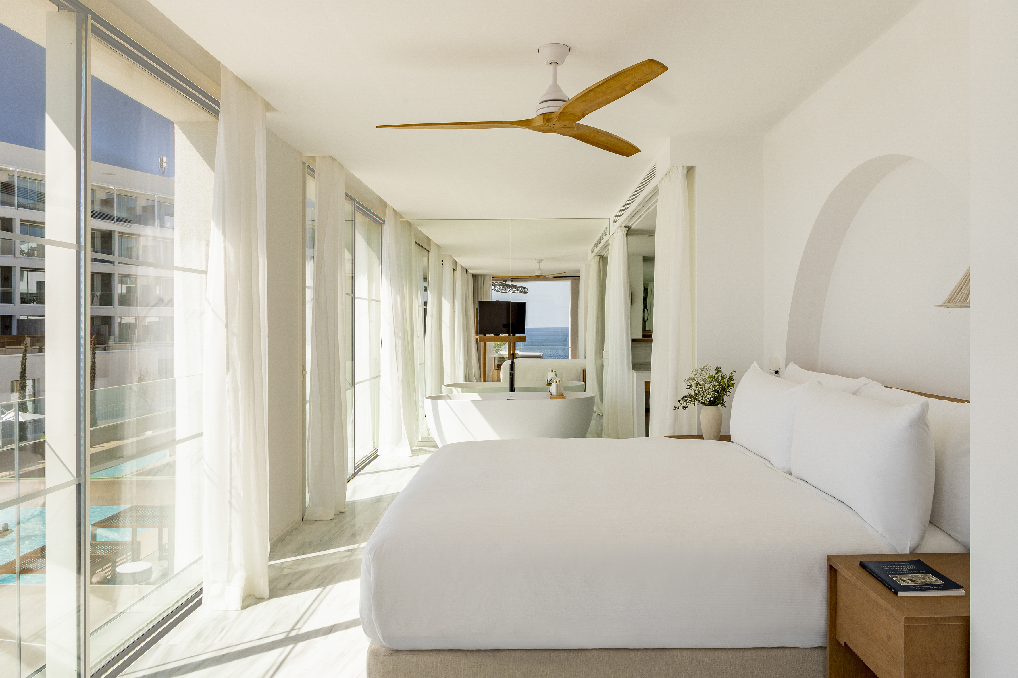 Bright white bedroom with ocean view, large windows, and freestanding bathtub.