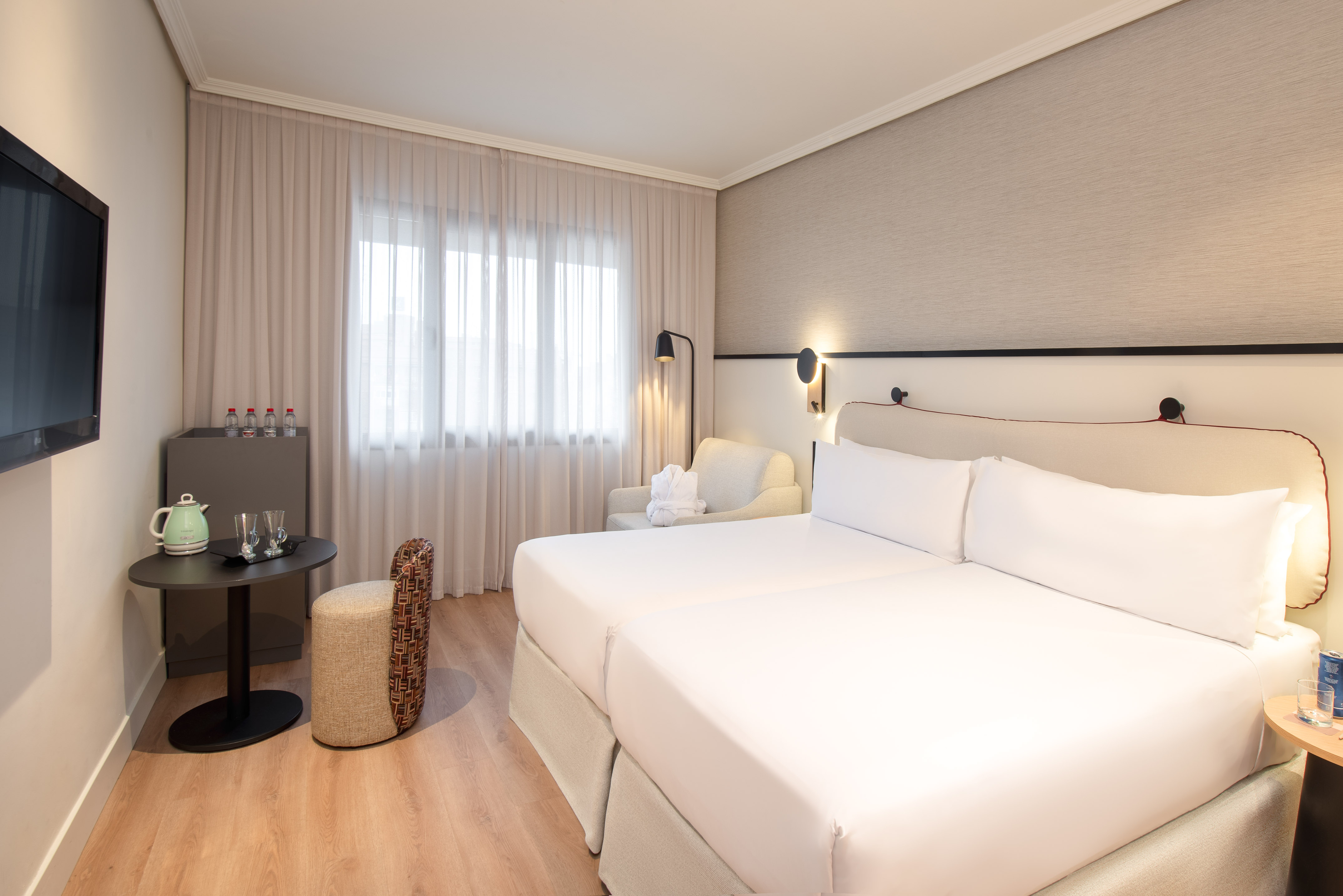 Bright hotel room with twin beds, armchair, TV, and window.
