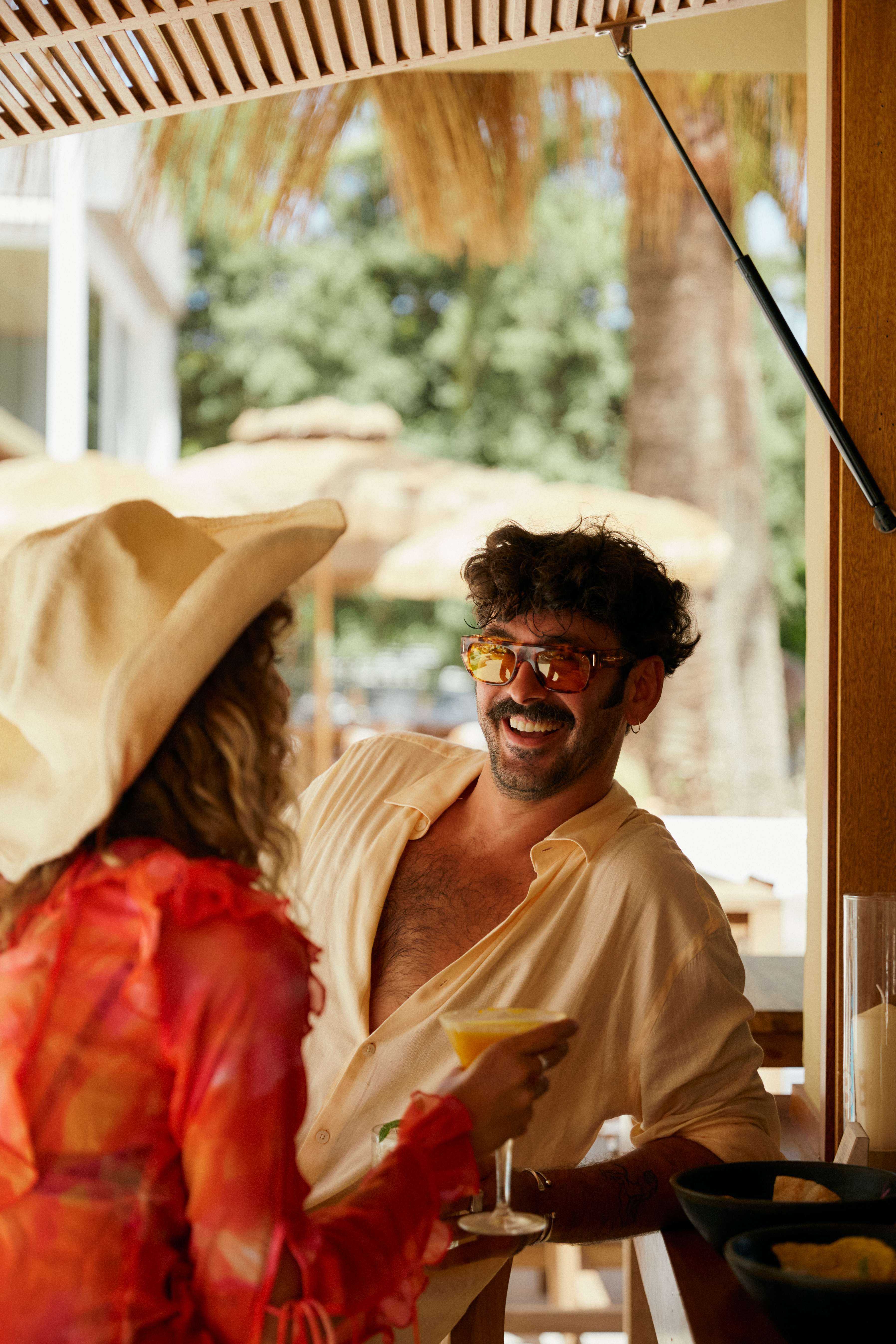 Man in sunglasses and woman in hat smiling with cocktails at a tropical bar.