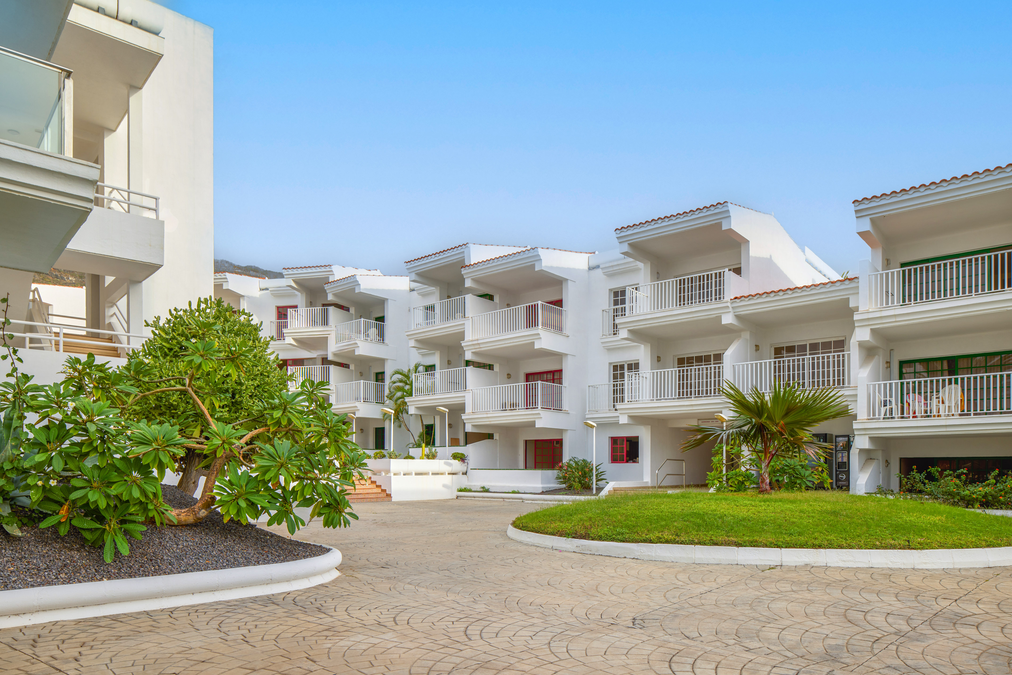 White resort apartments with balconies, tropical landscaping, and sunny blue sky.