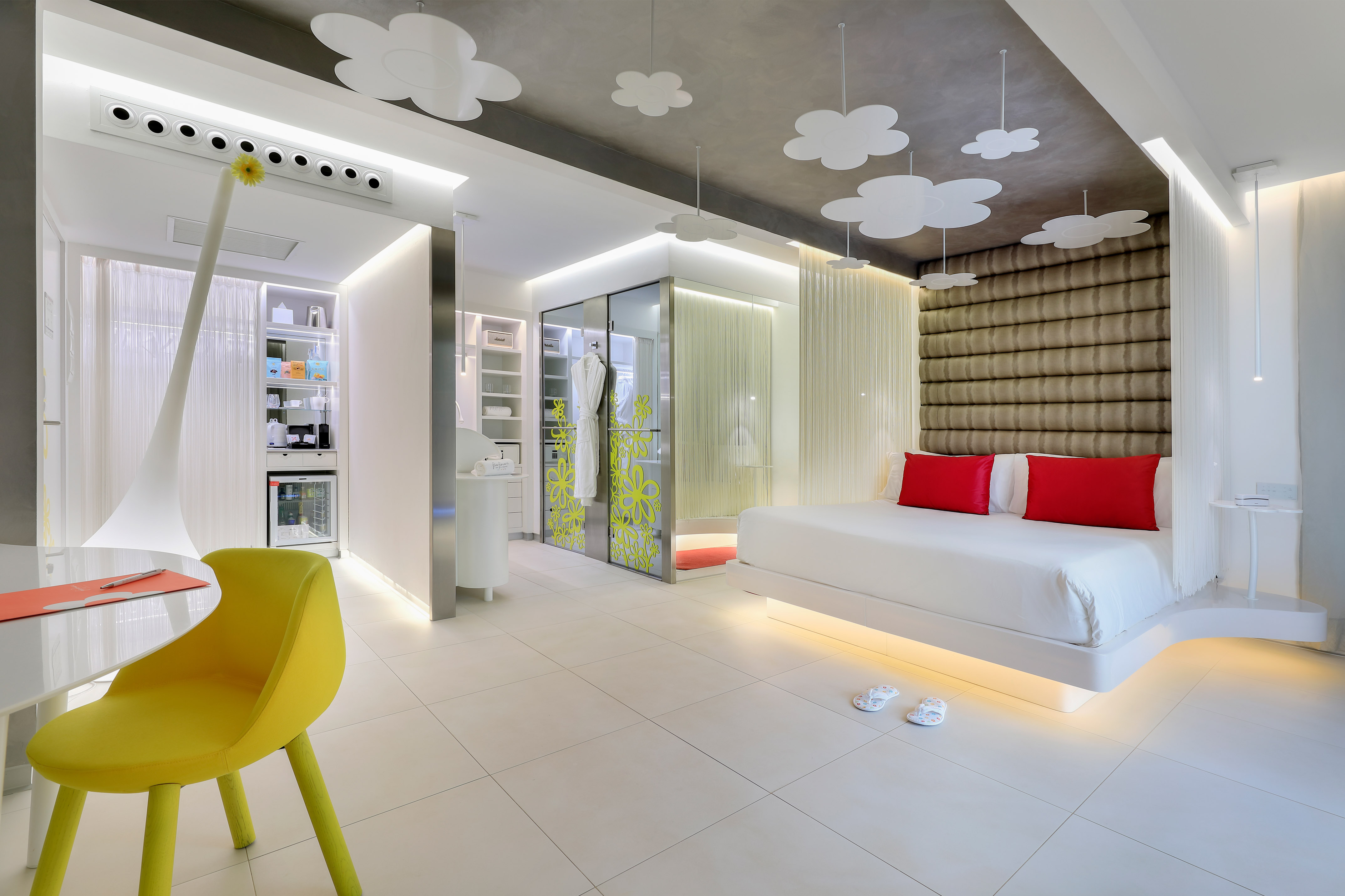 Modern hotel room: floating bed, red pillows, yellow chair, flower lights.