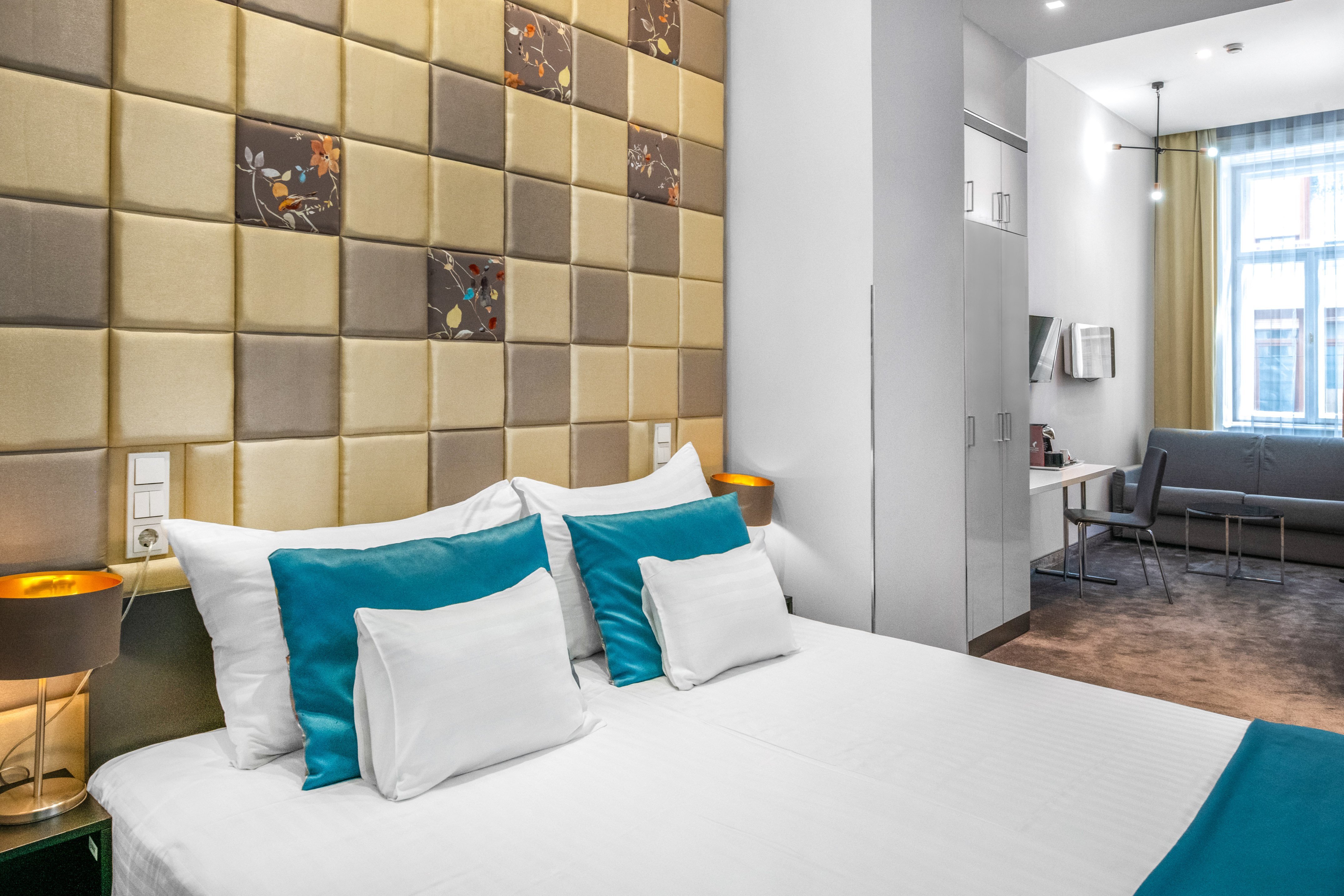 Hotel bedroom with white bed, teal pillows, and patterned headboard.
