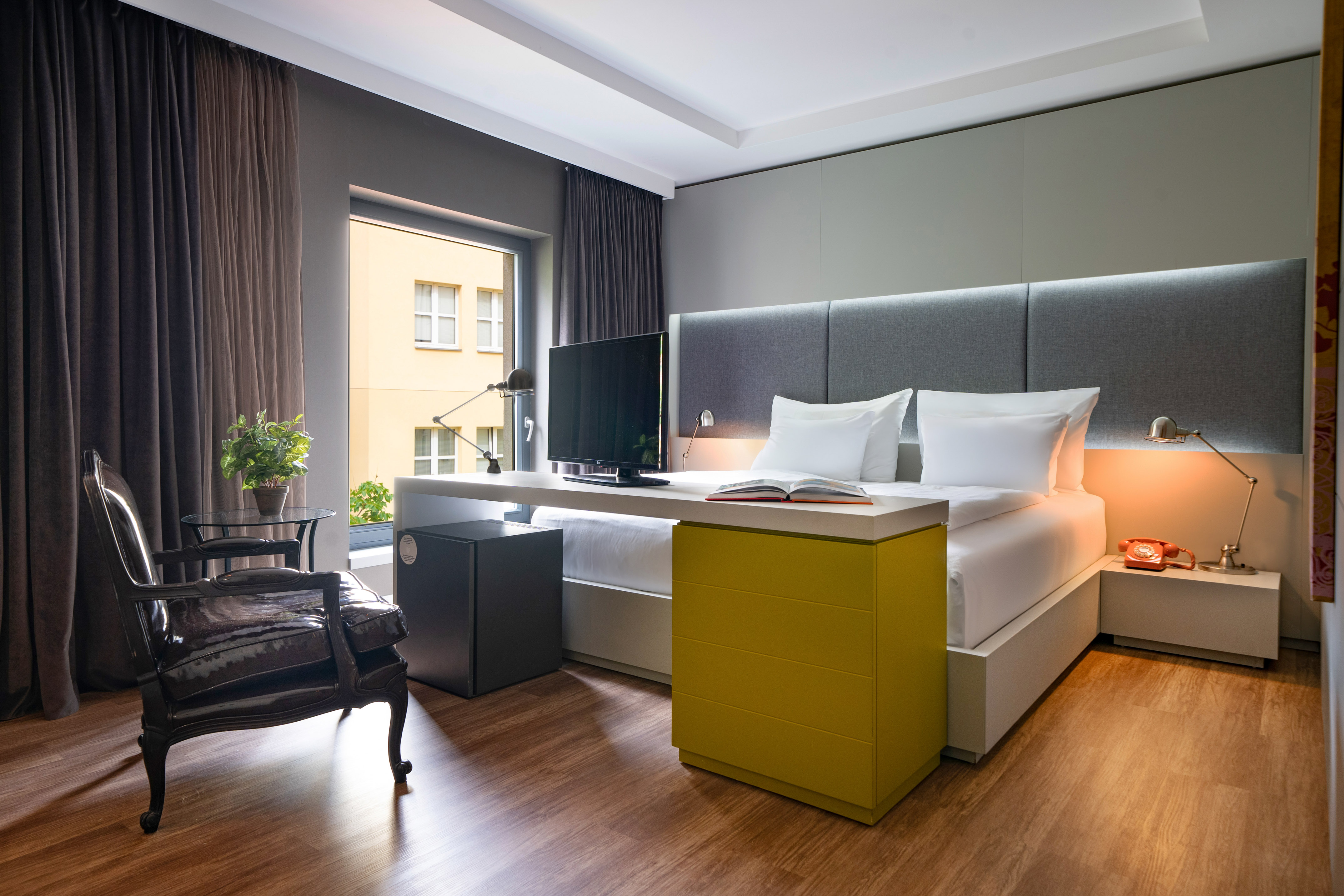 Modern hotel bedroom with large bed, TV, desk, yellow cabinet, and brown curtains.