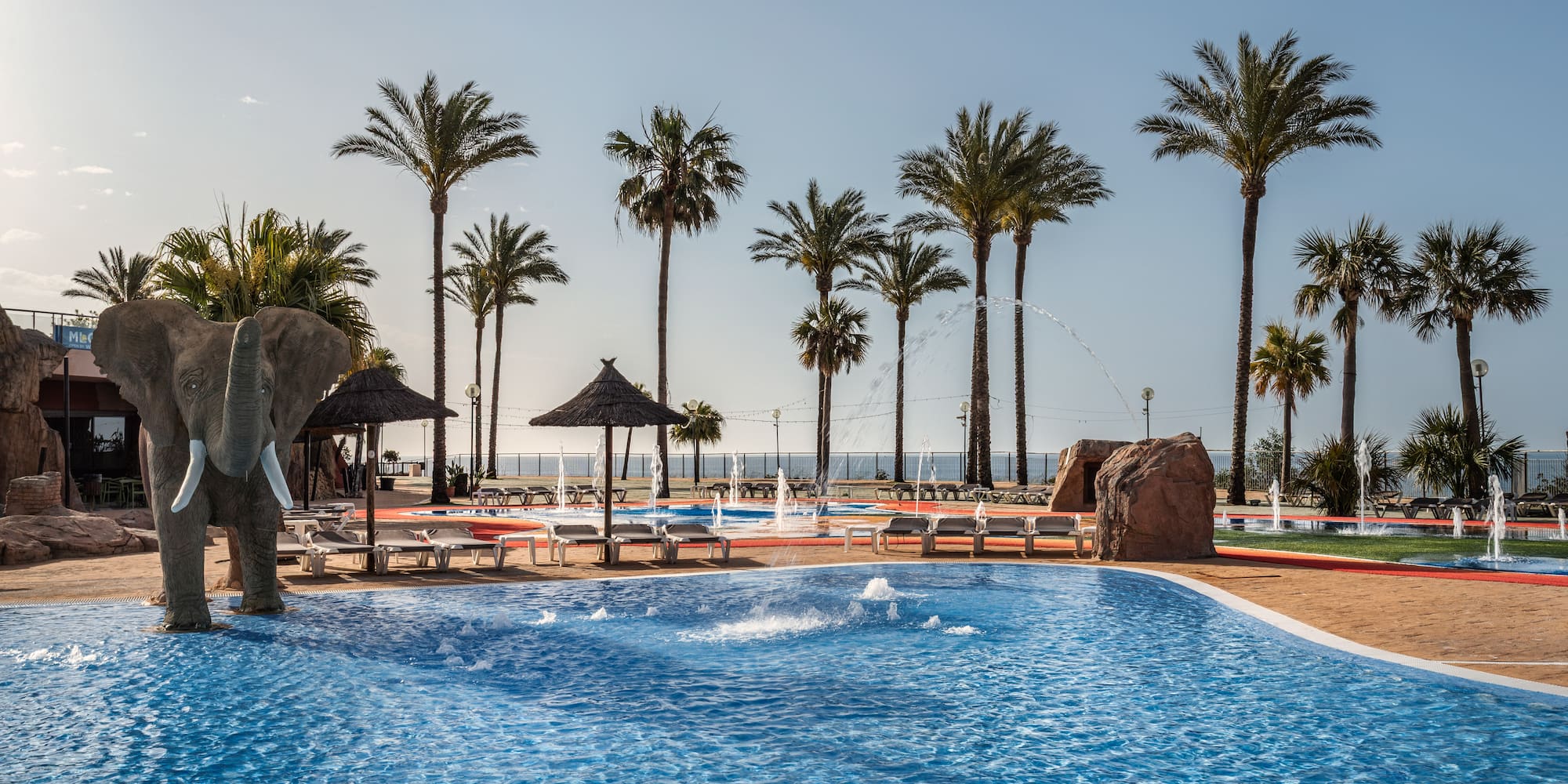 Tropical resort pool with elephant statue, palm trees, and water fountains.