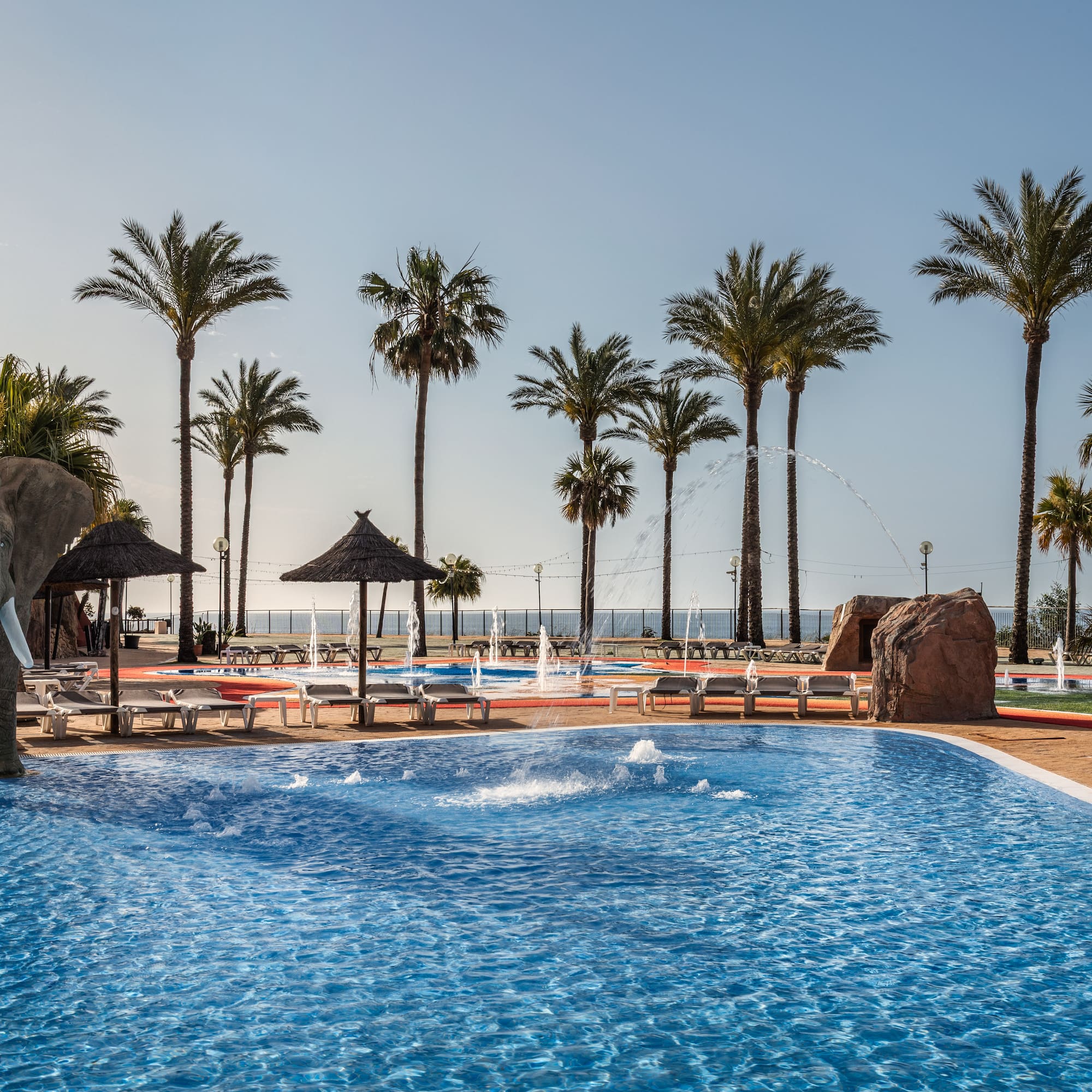 Resort swimming pool with elephant statue, palm trees, fountains, and sun loungers.