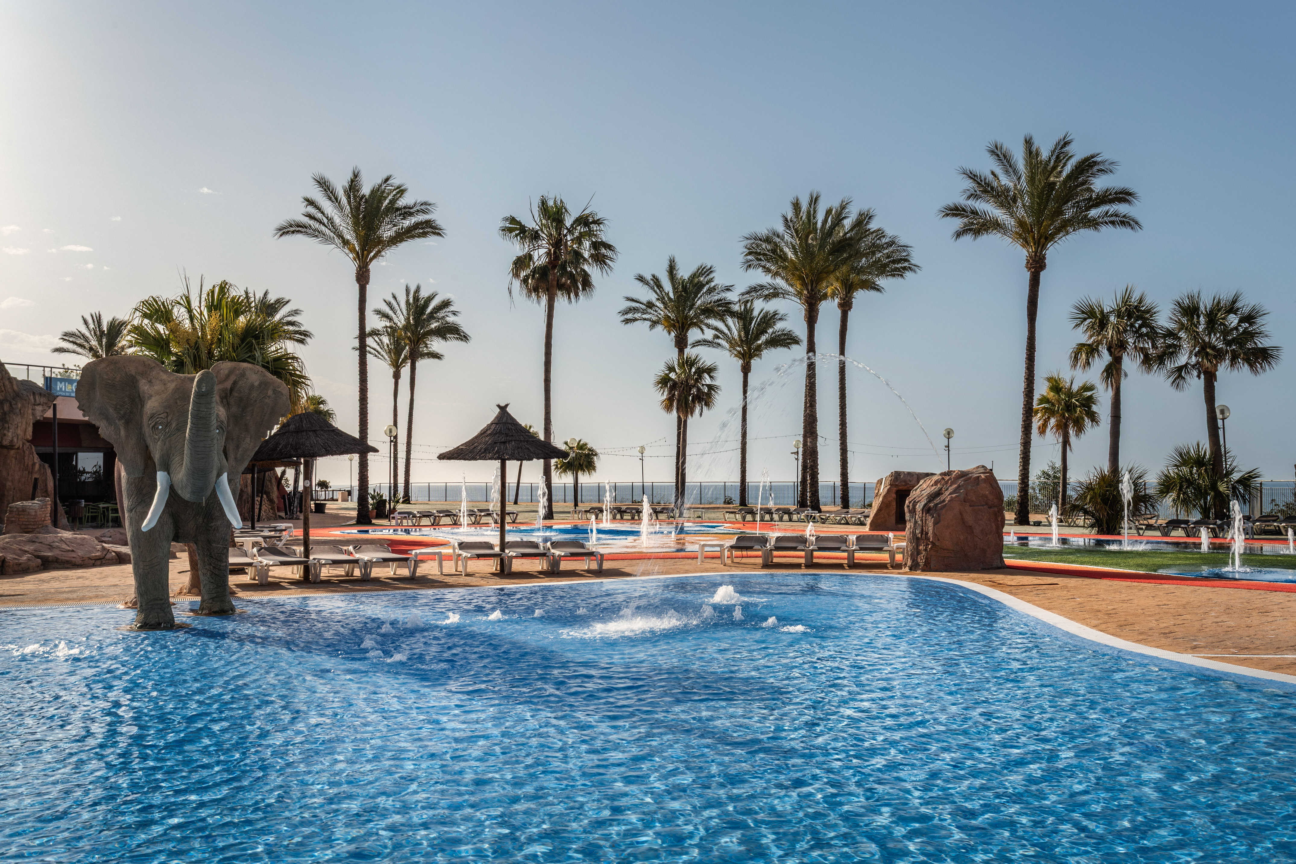 Tropical resort pool with elephant statue, palm trees, and water fountains.