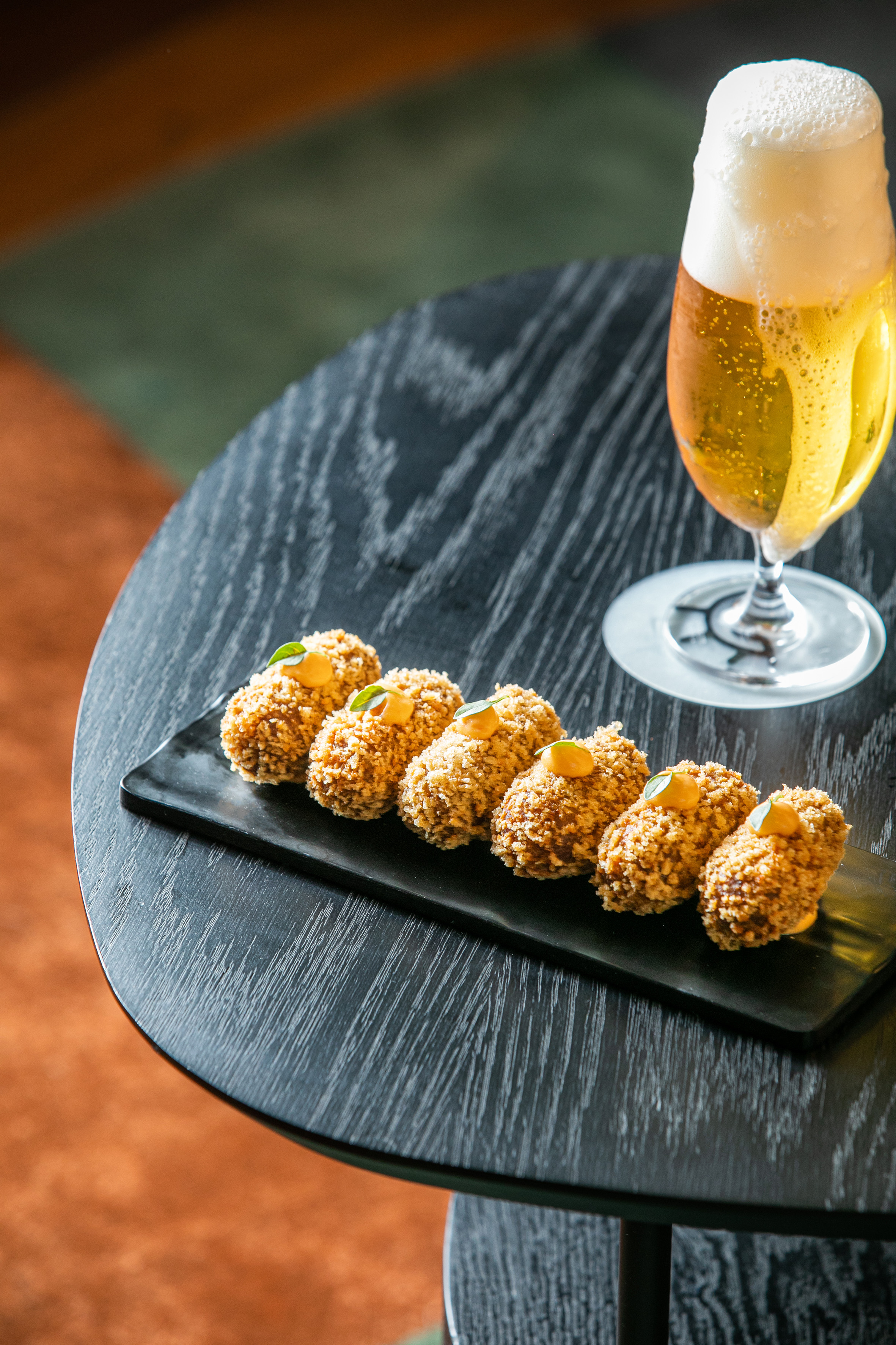 Crispy croquettes with orange sauce, green garnish, and beer on a dark table.