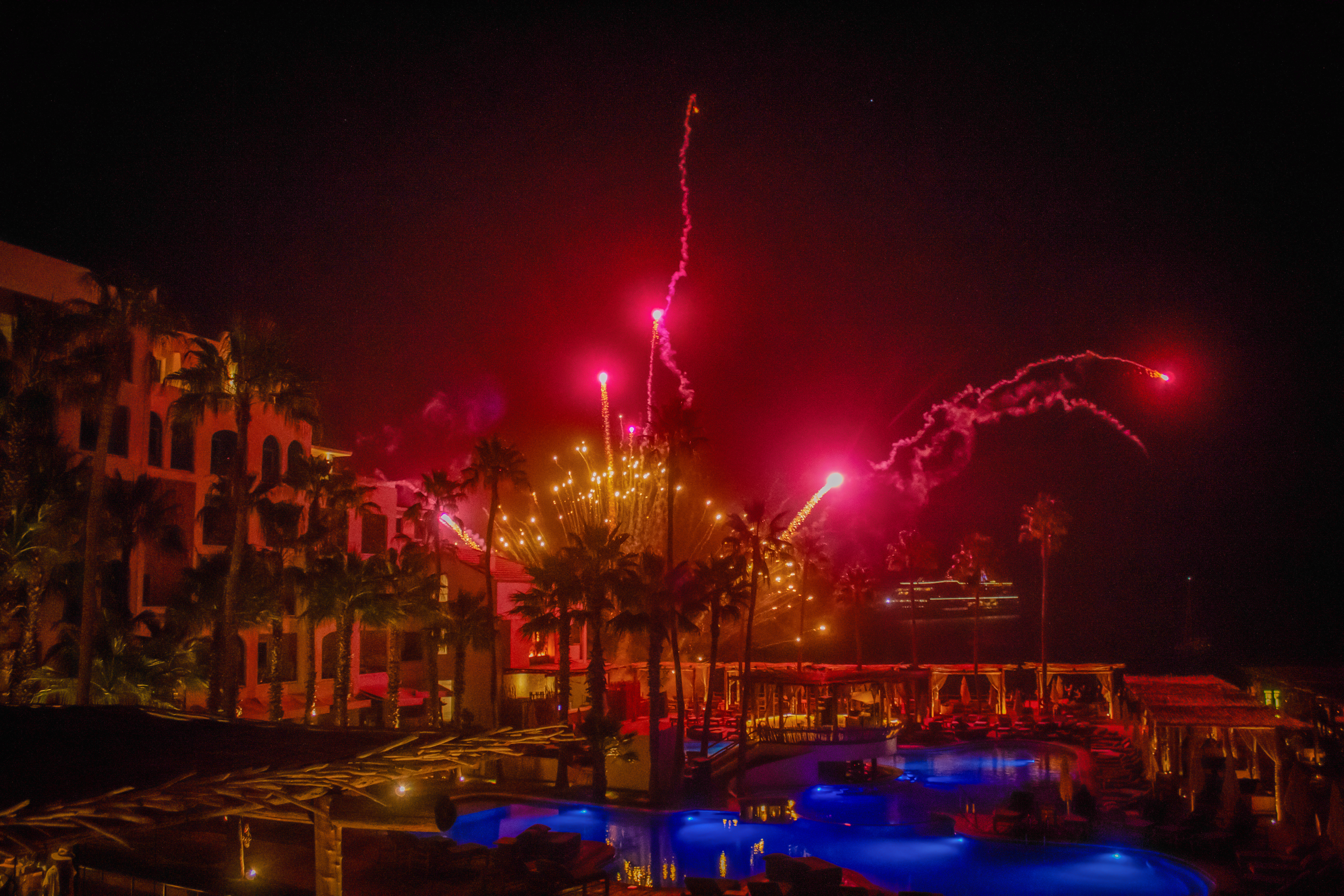 Night fireworks display over a luxury resort with palm trees and blue lit pool.