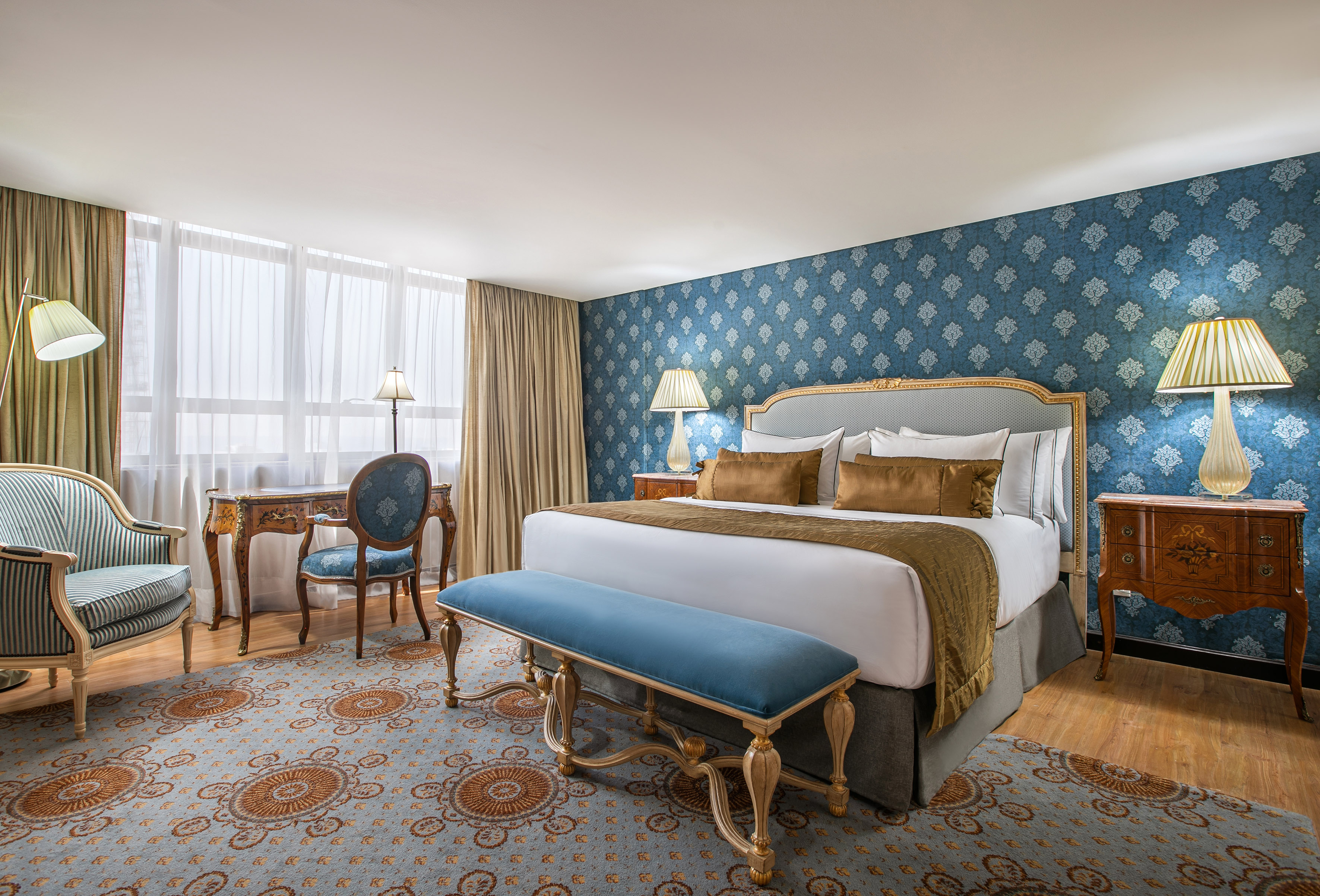Luxury hotel bedroom with blue patterned wallpaper, king bed, and ornate furniture.