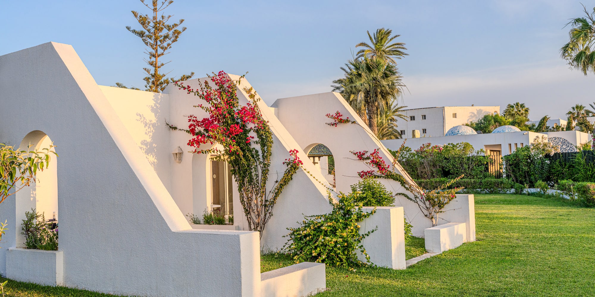 Mediterranean white building with bougainvillea, palm trees, and green lawn.
