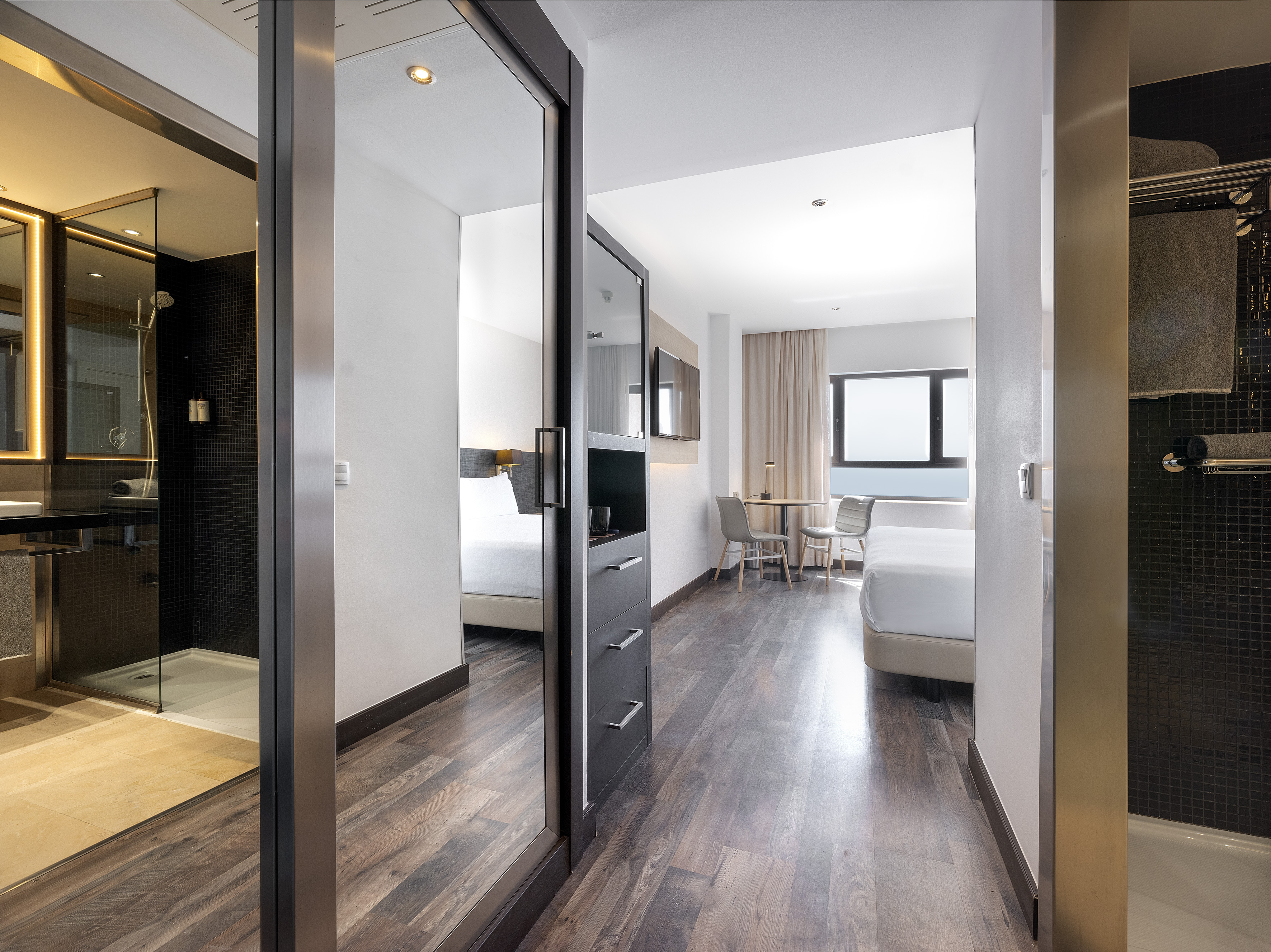 Modern hotel room with glass-partitioned bathroom, bedroom, and wood floor.