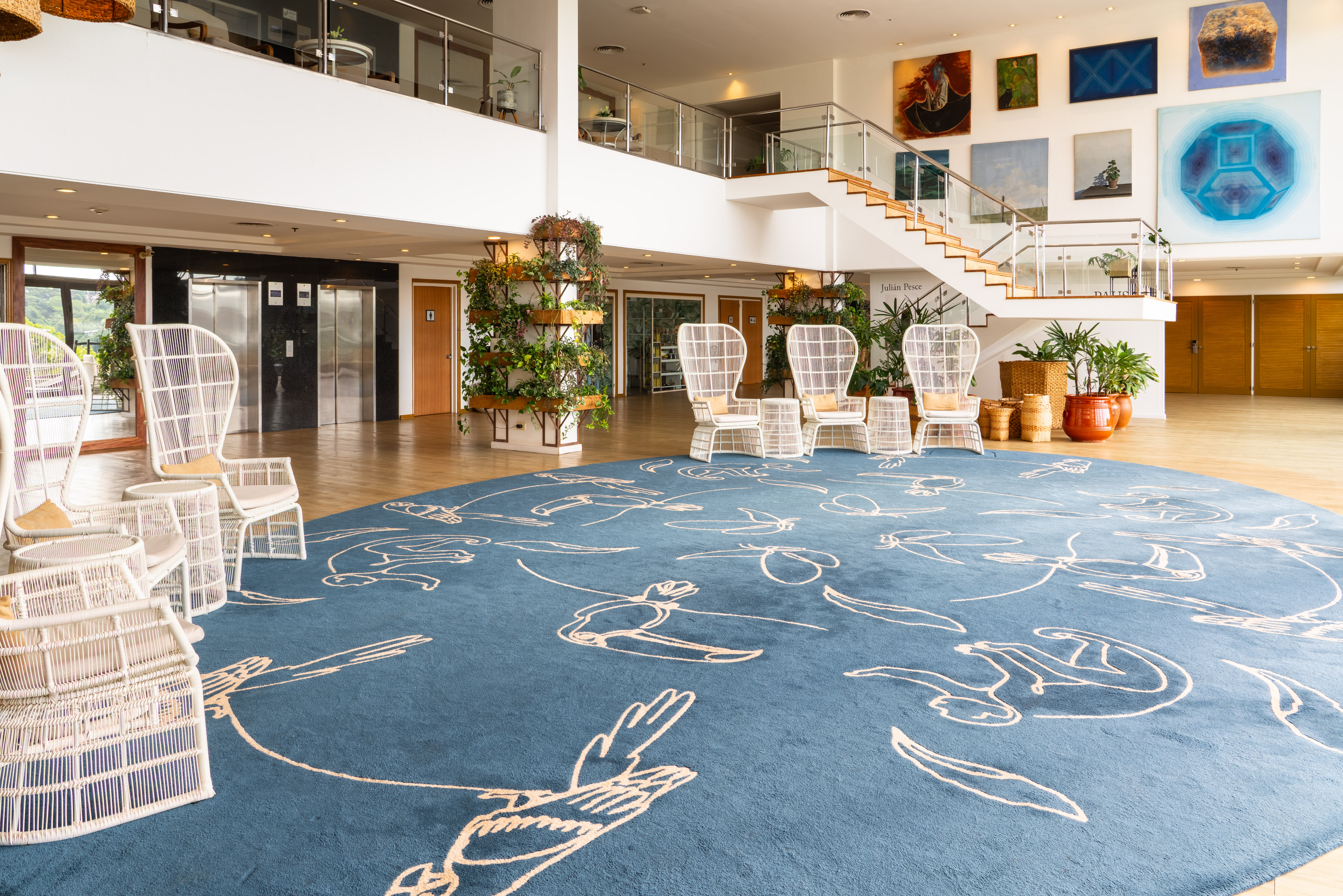 Elegant hotel lobby, blue rug, white wicker chairs, grand staircase, diverse artworks.