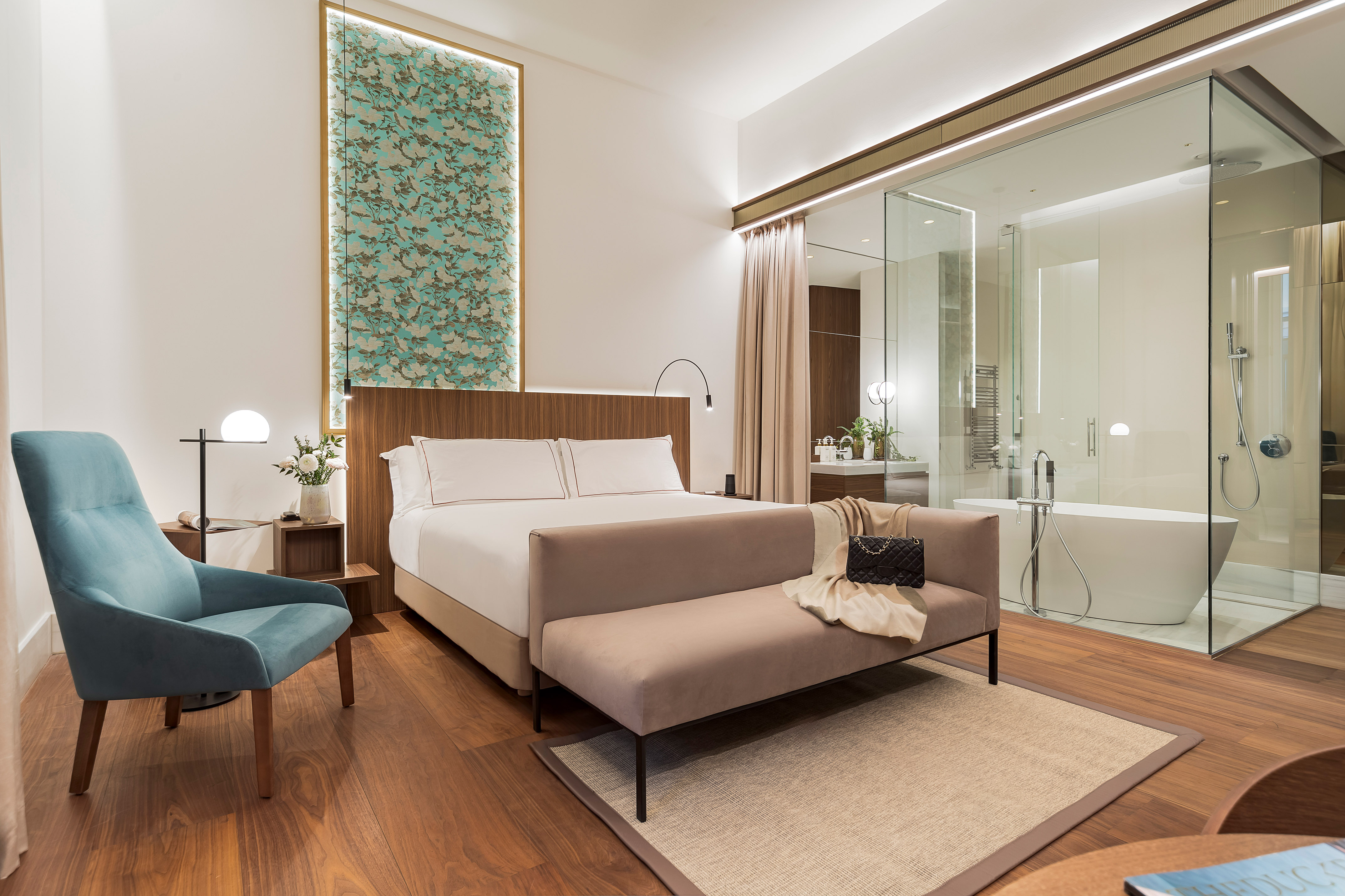 Modern hotel bedroom with glass bathroom, queen bed, and blue accent chair.
