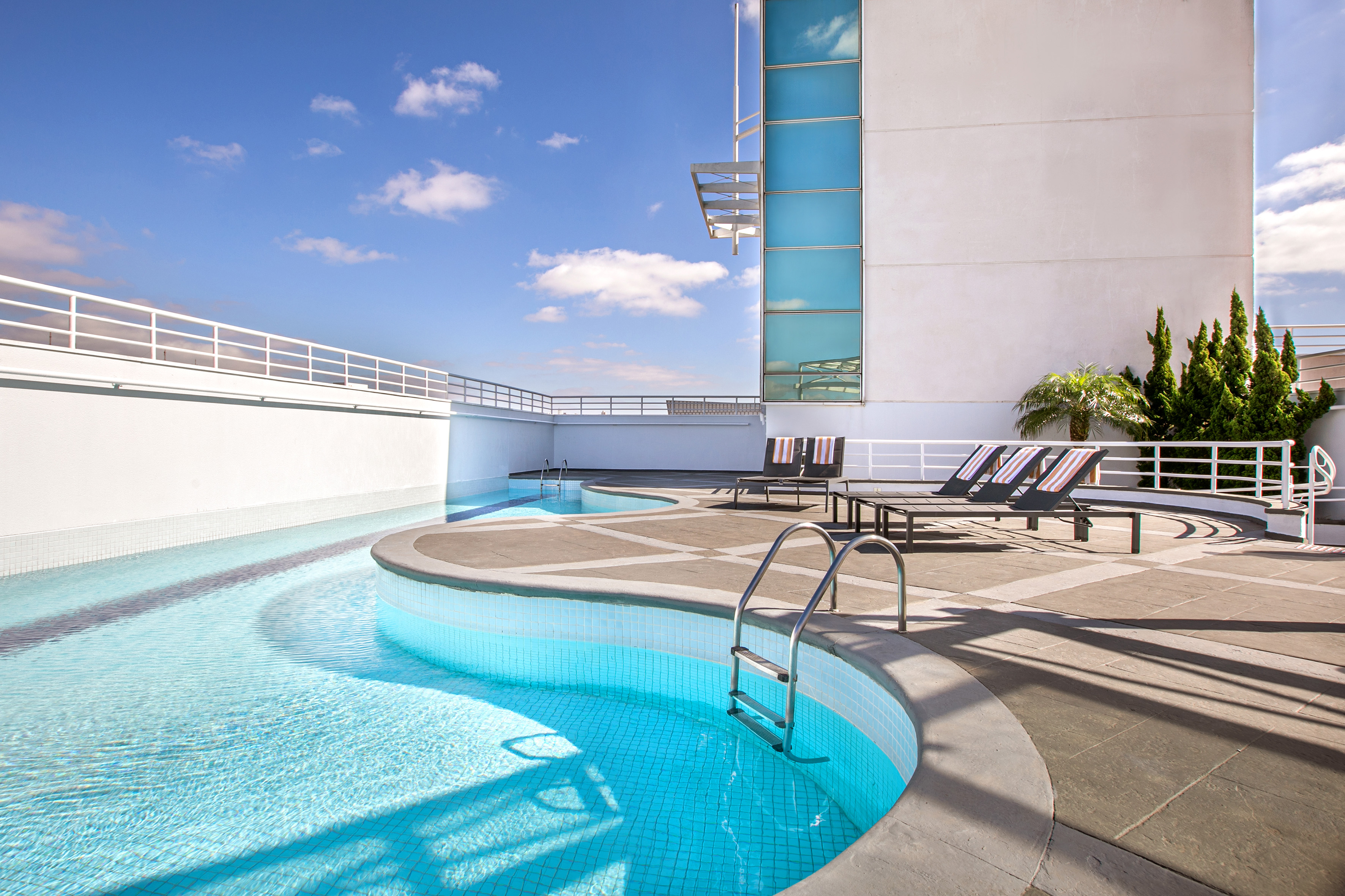 Modern hotel rooftop pool with lounge chairs under a clear blue sky.