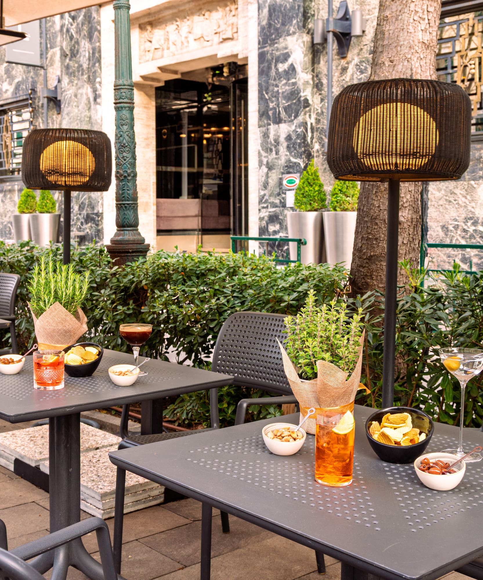 Outdoor patio cafe with cocktails, snacks, dark tables, and illuminated lamps.