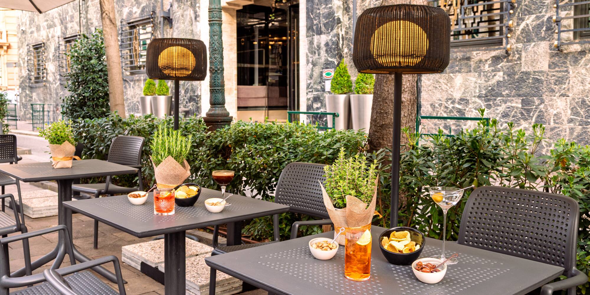 Outdoor patio cafe with cocktails, snacks, dark tables, and illuminated lamps.