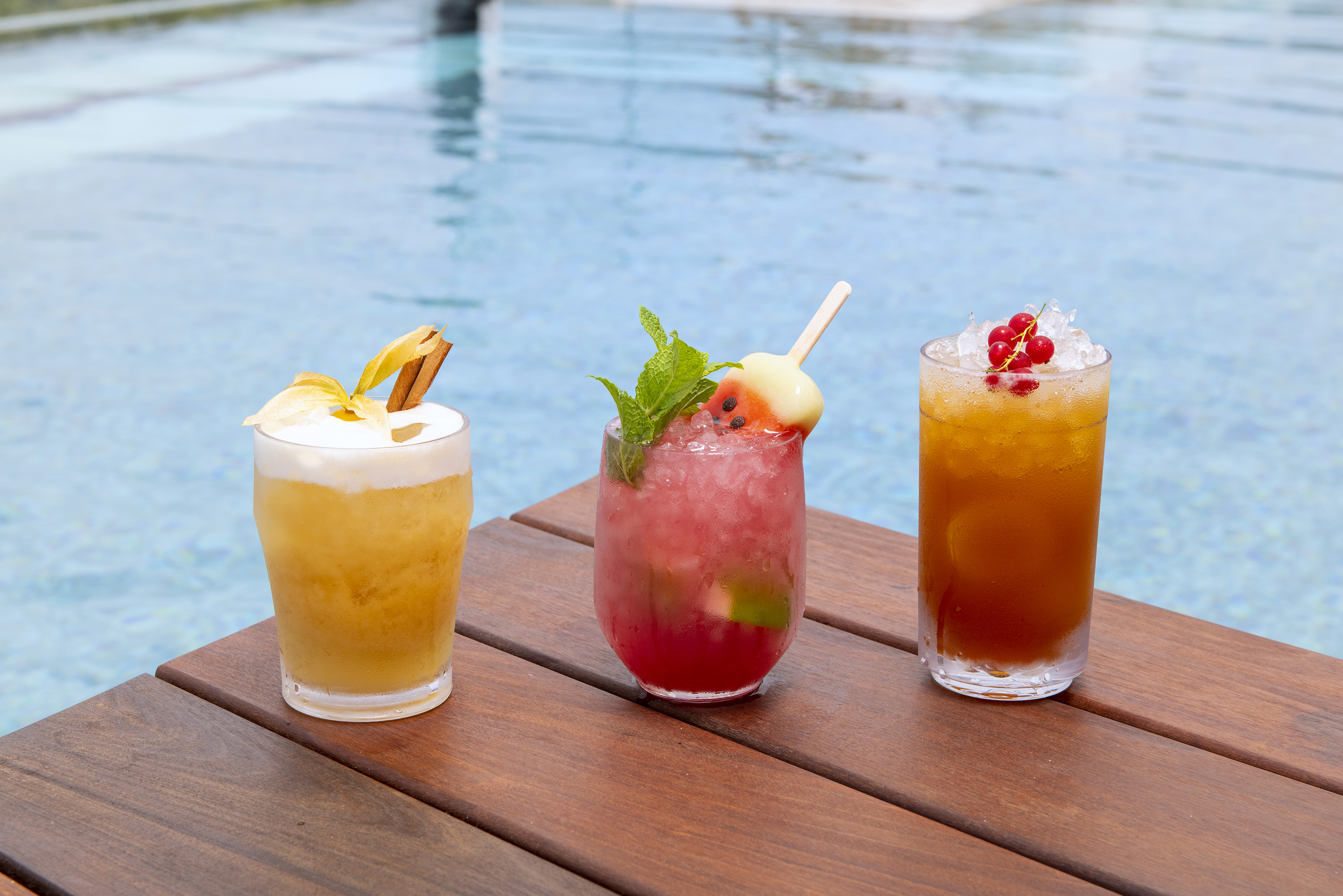 Three refreshing cocktails on a wooden table by a blue pool.