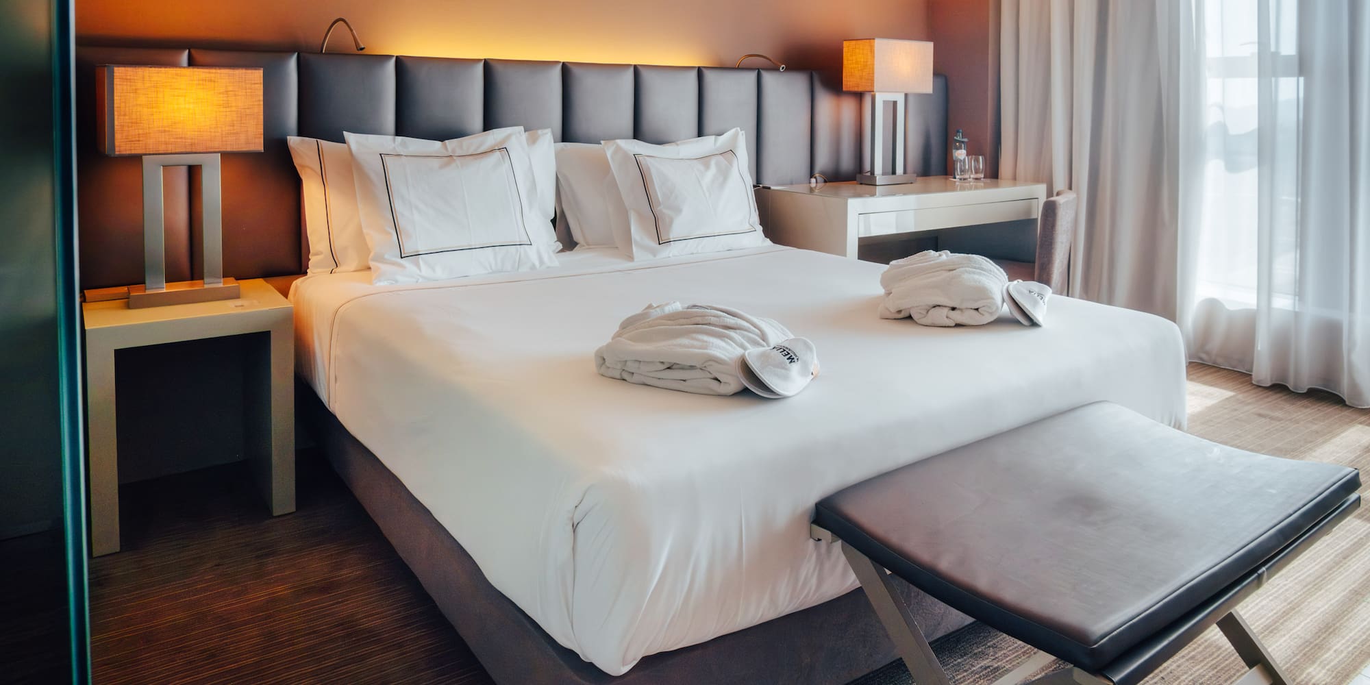 Modern hotel room with bed, white linens, bathrobes, lamps, and sheer curtains.