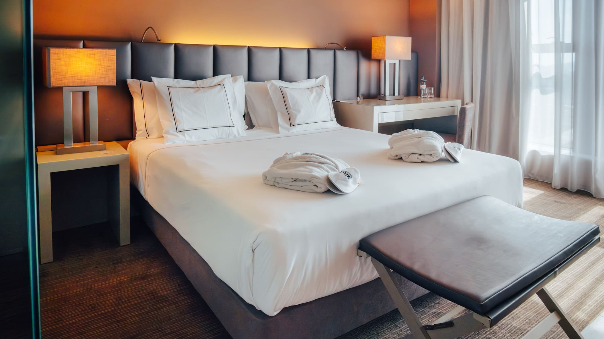 Modern hotel room with bed, white linens, bathrobes, lamps, and sheer curtains.