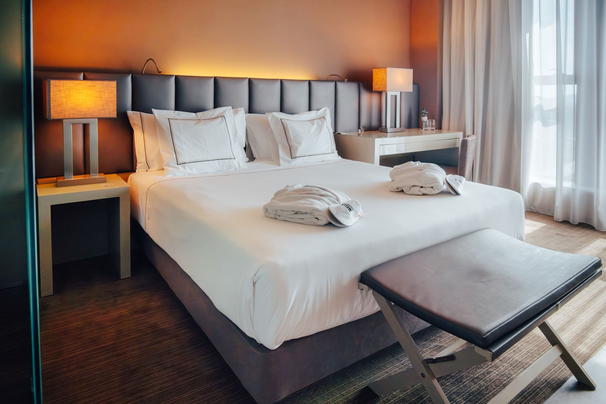 Modern hotel room with bed, white linens, bathrobes, lamps, and sheer curtains.